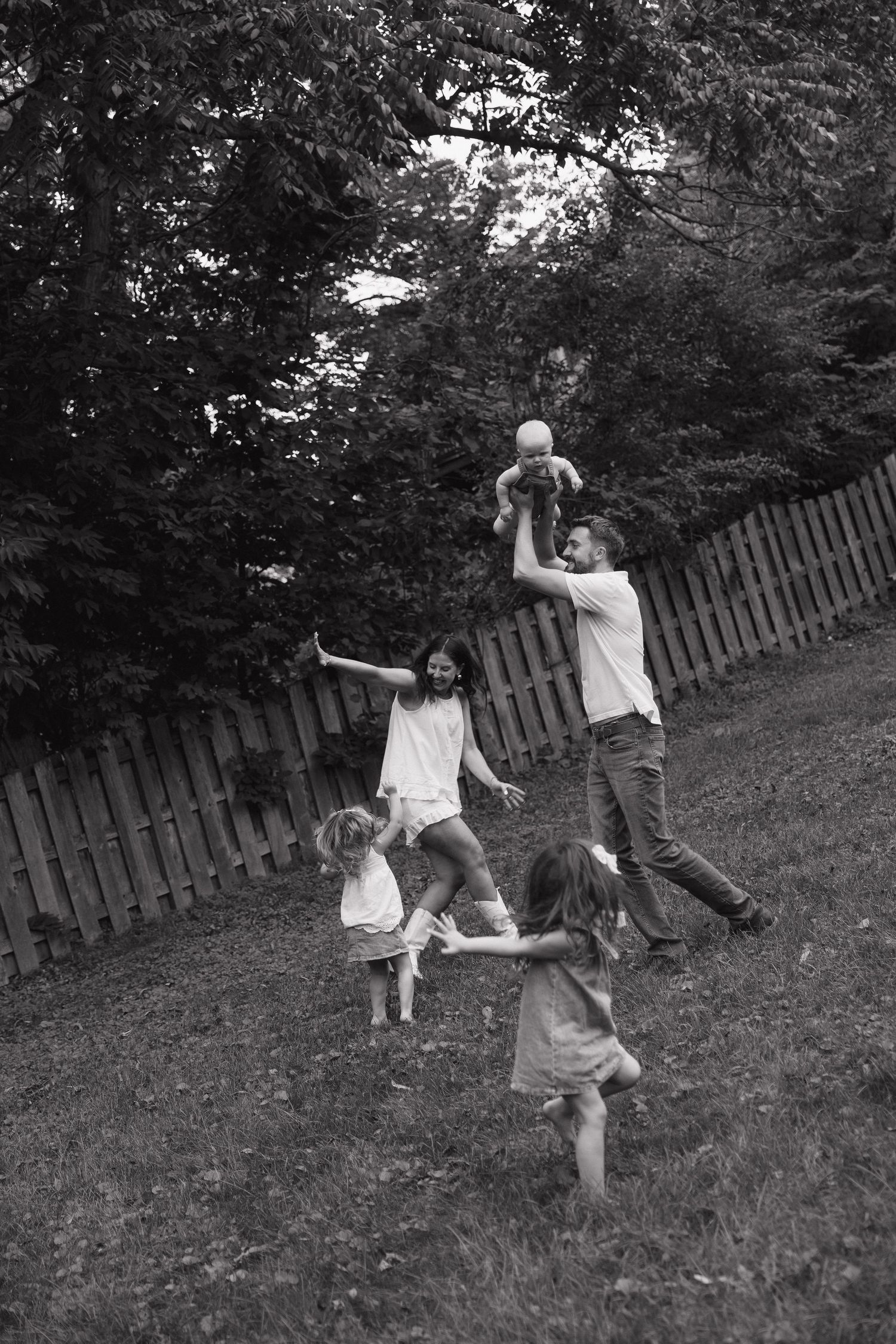 Black and white photo of a joyful family playing outdoors, father lifting baby while children run on grass near wooden fence.
