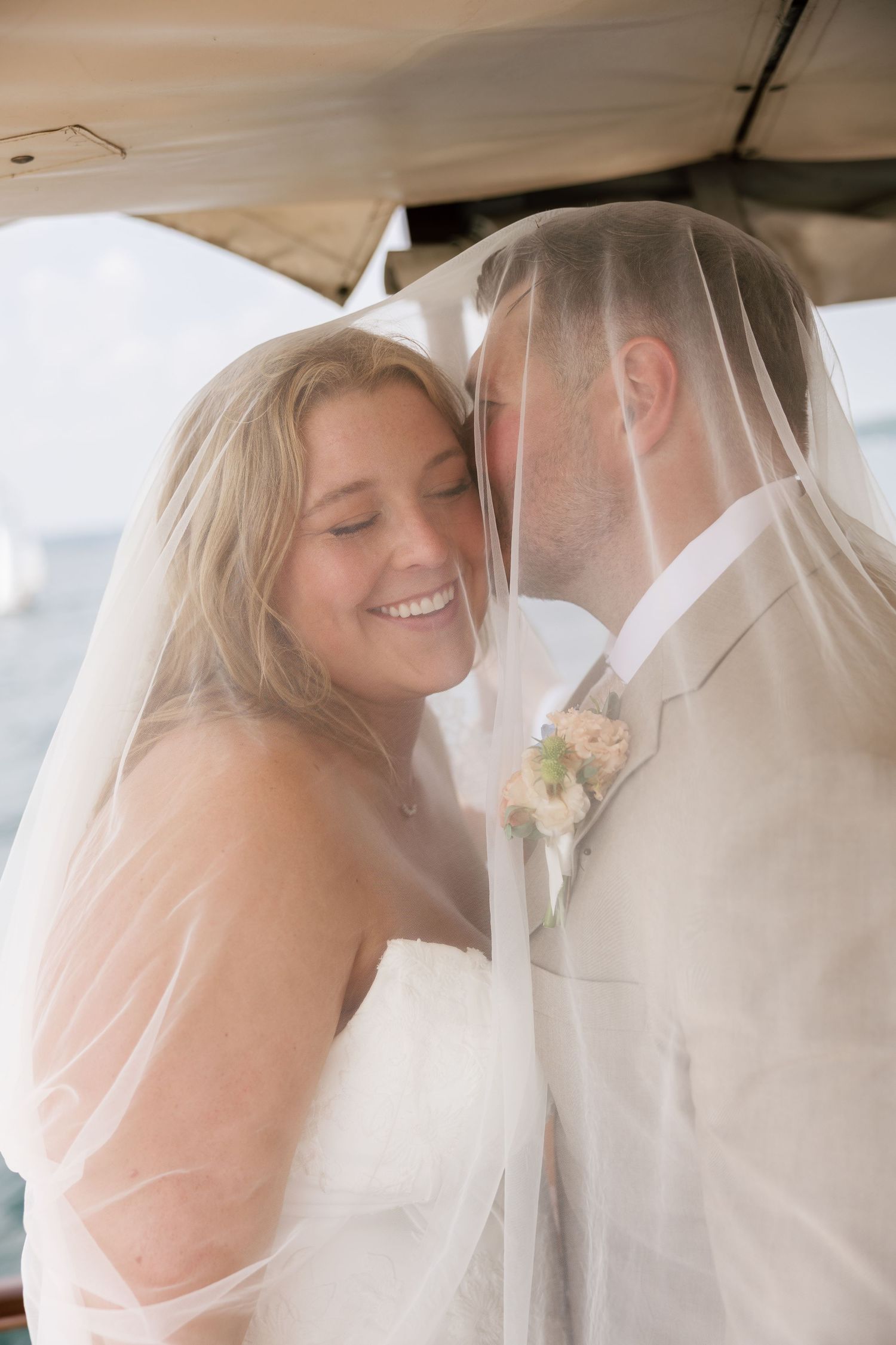 Bride and groom share a romantic moment under a wedding veil on a boat, bride smiling joyfully.