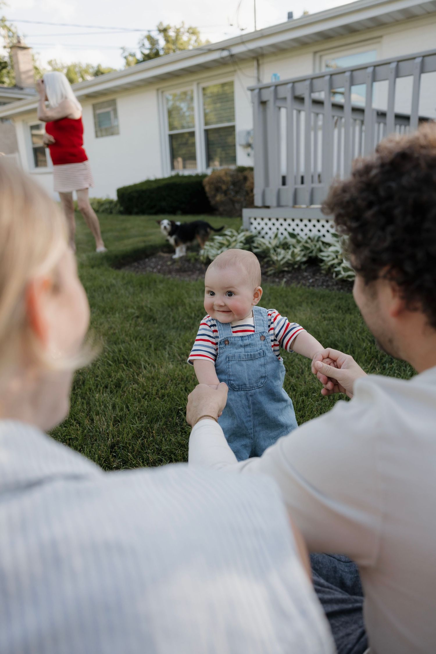 Baby in denim overalls smiles at parents sitting on grass in a sunny backyard with a dog nearby.