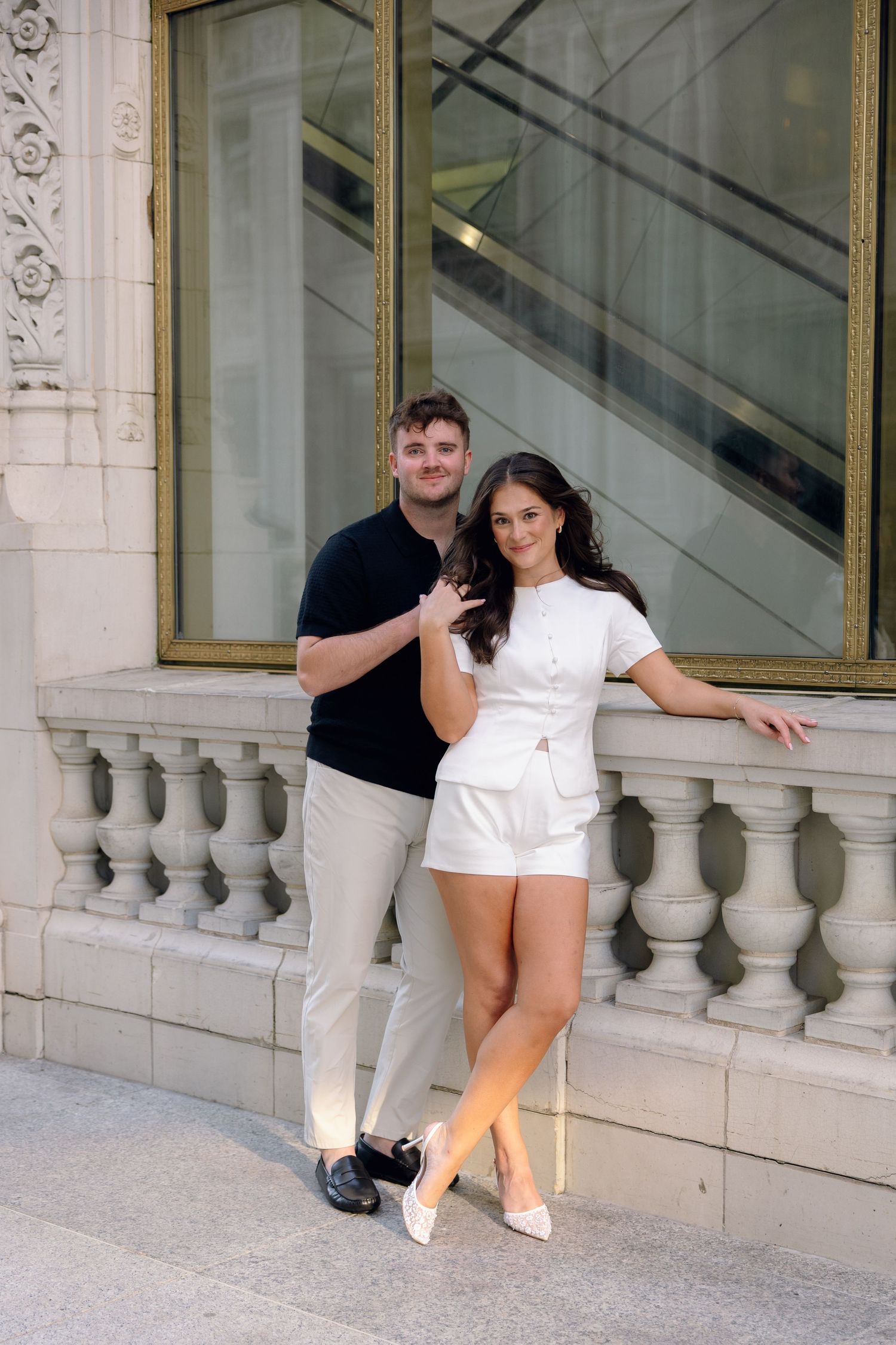 A couple poses by ornate white stone balustrade, man in black shirt and khakis, woman in white romper and heels.