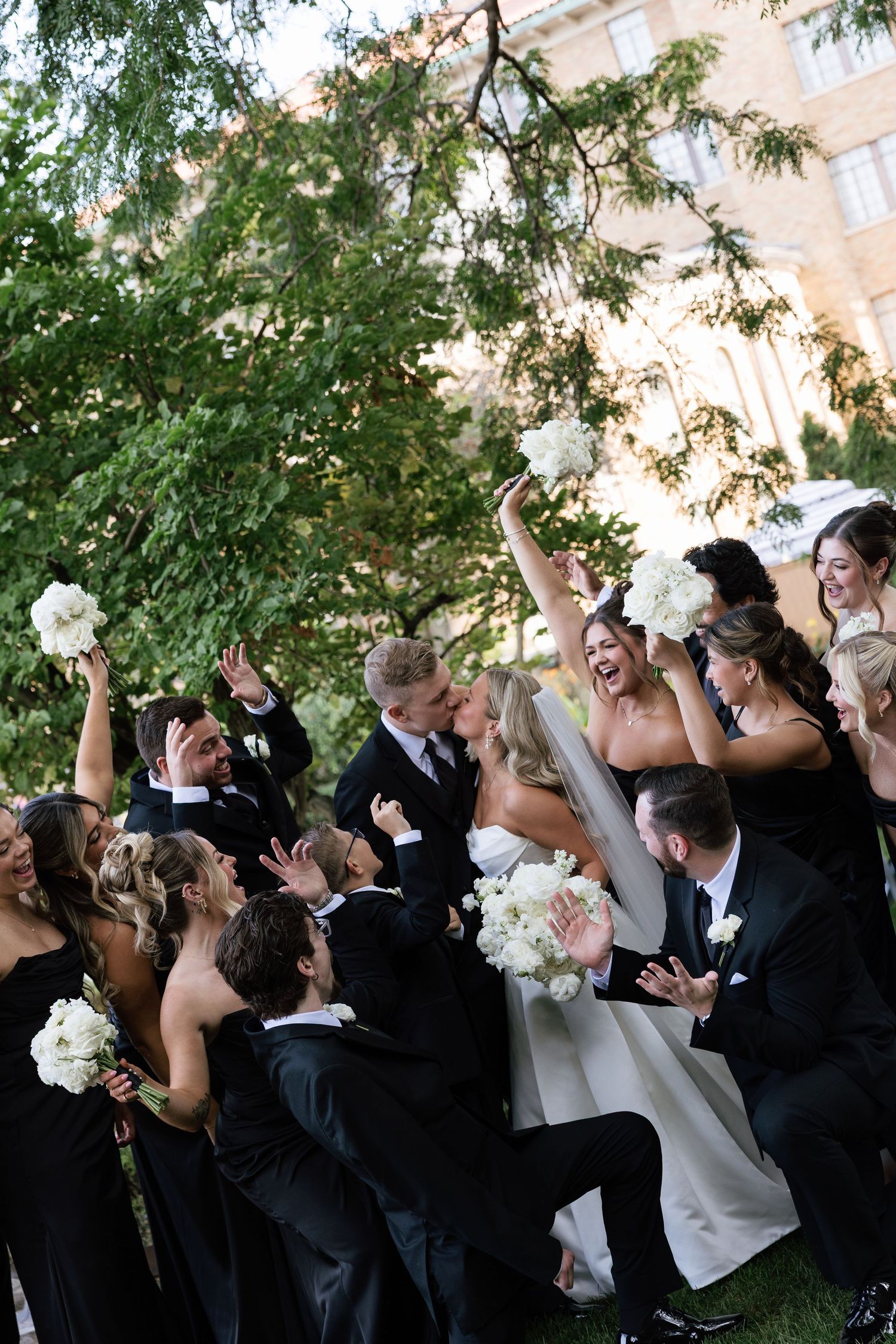 Wedding party in black attire cheers as bride and groom kiss outdoors near lush greenery and elegant building.
