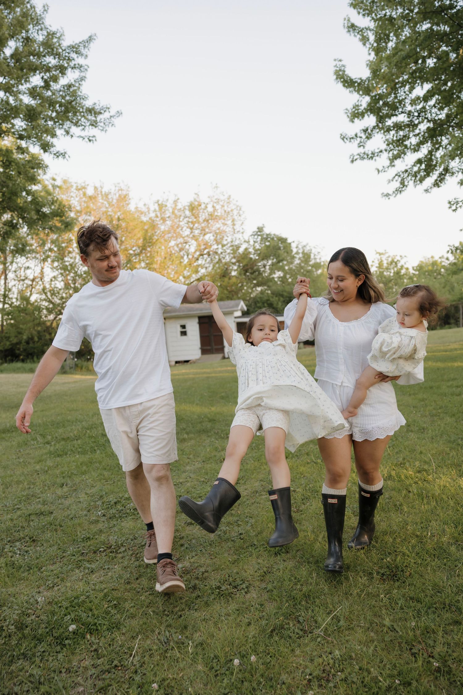 Happy family in white outfits swinging child between parents on green lawn with trees.