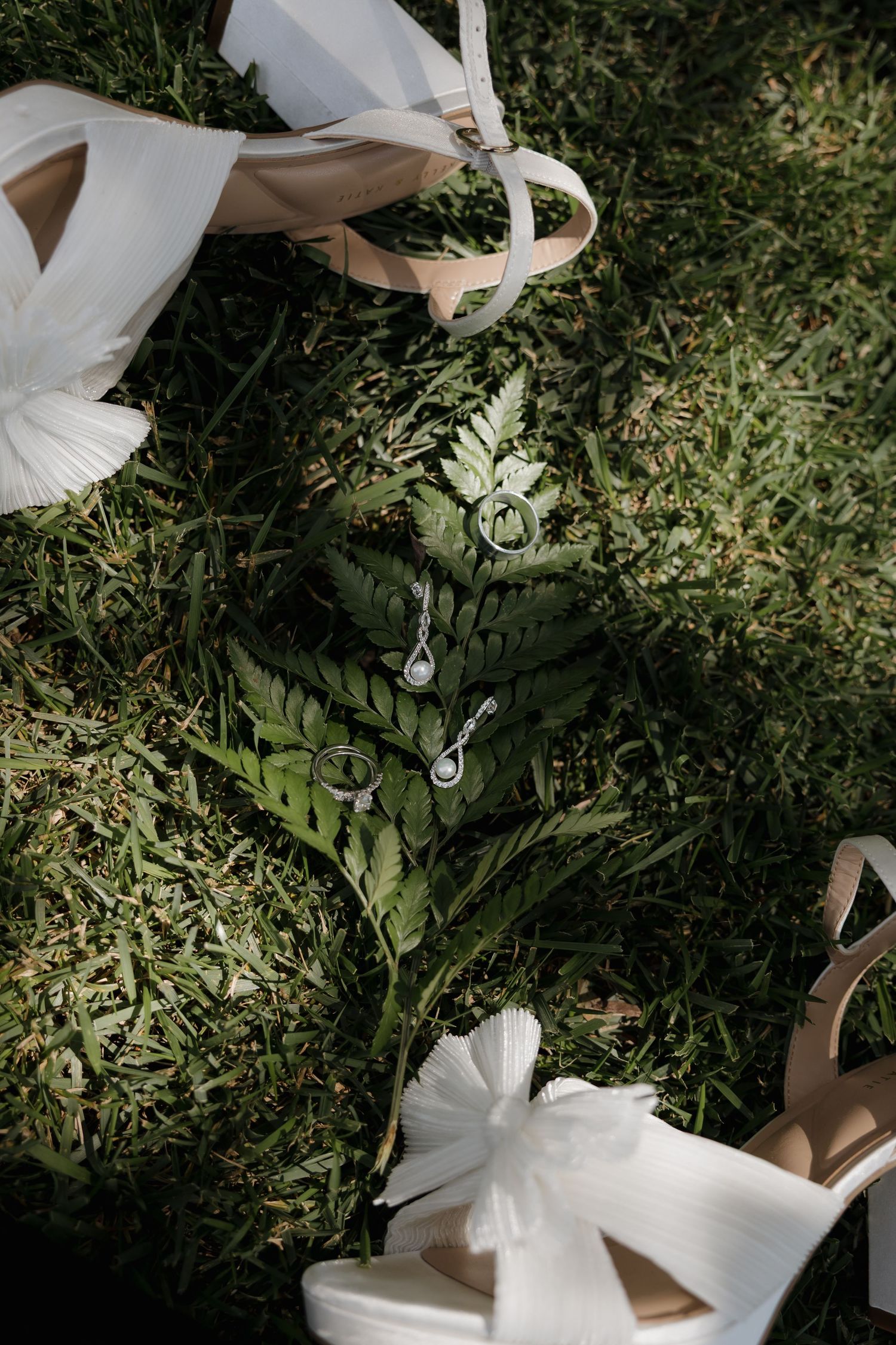 Wedding rings and earrings displayed on a green fern leaf surrounded by bridal heels on grass.