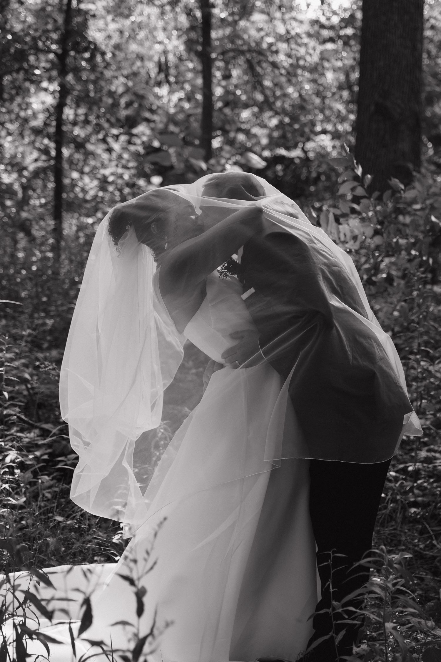 Bride and groom sharing a romantic kiss under the wedding veil in a lush forest setting, black and white photo.