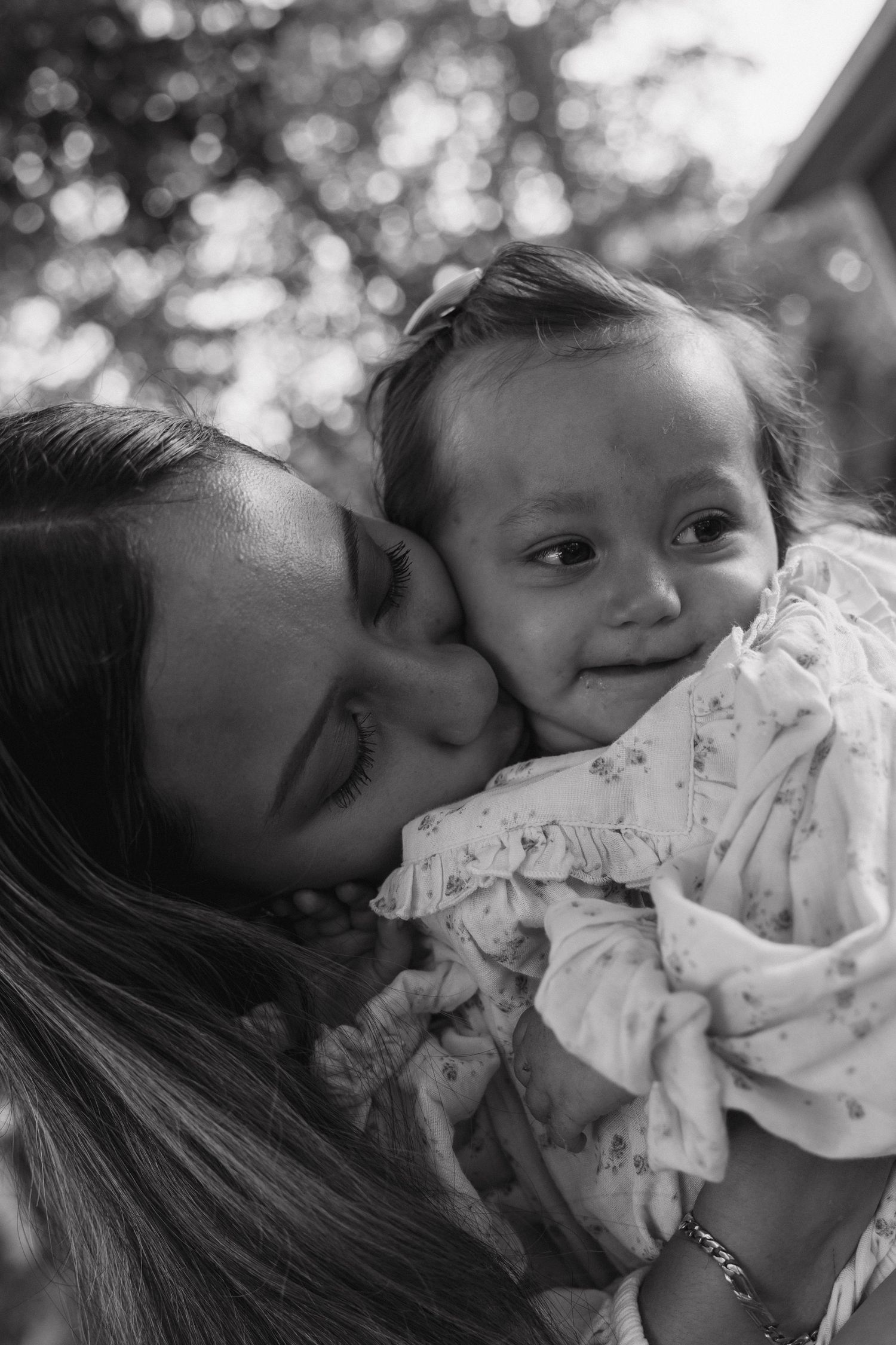 Black and white close-up of a mother tenderly kissing her smiling baby outdoors.
