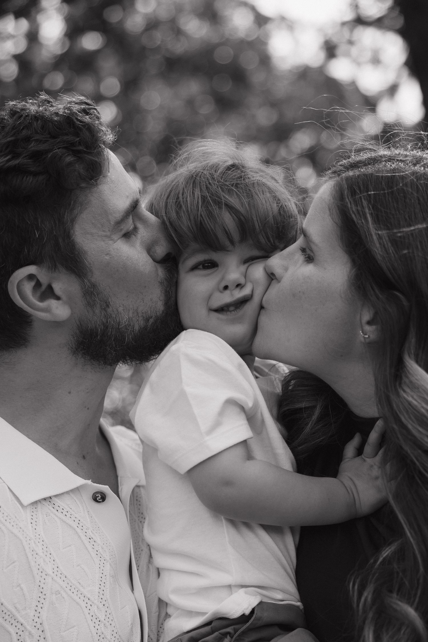 Black and white photo of parents kissing their happy toddler son on both cheeks.