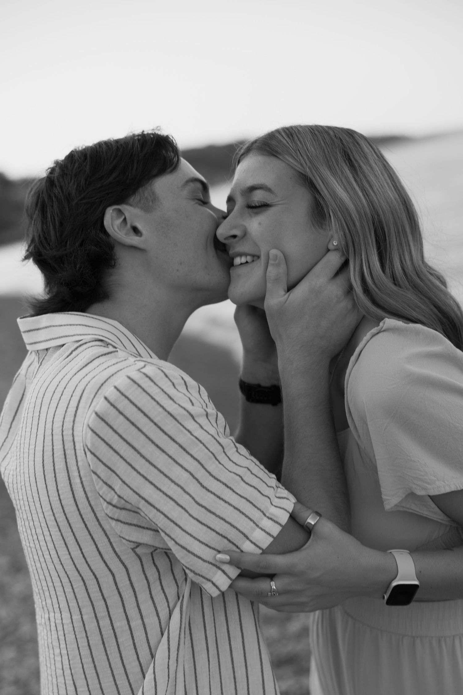 Black and white photo of a young couple sharing a romantic kiss outdoors near water.