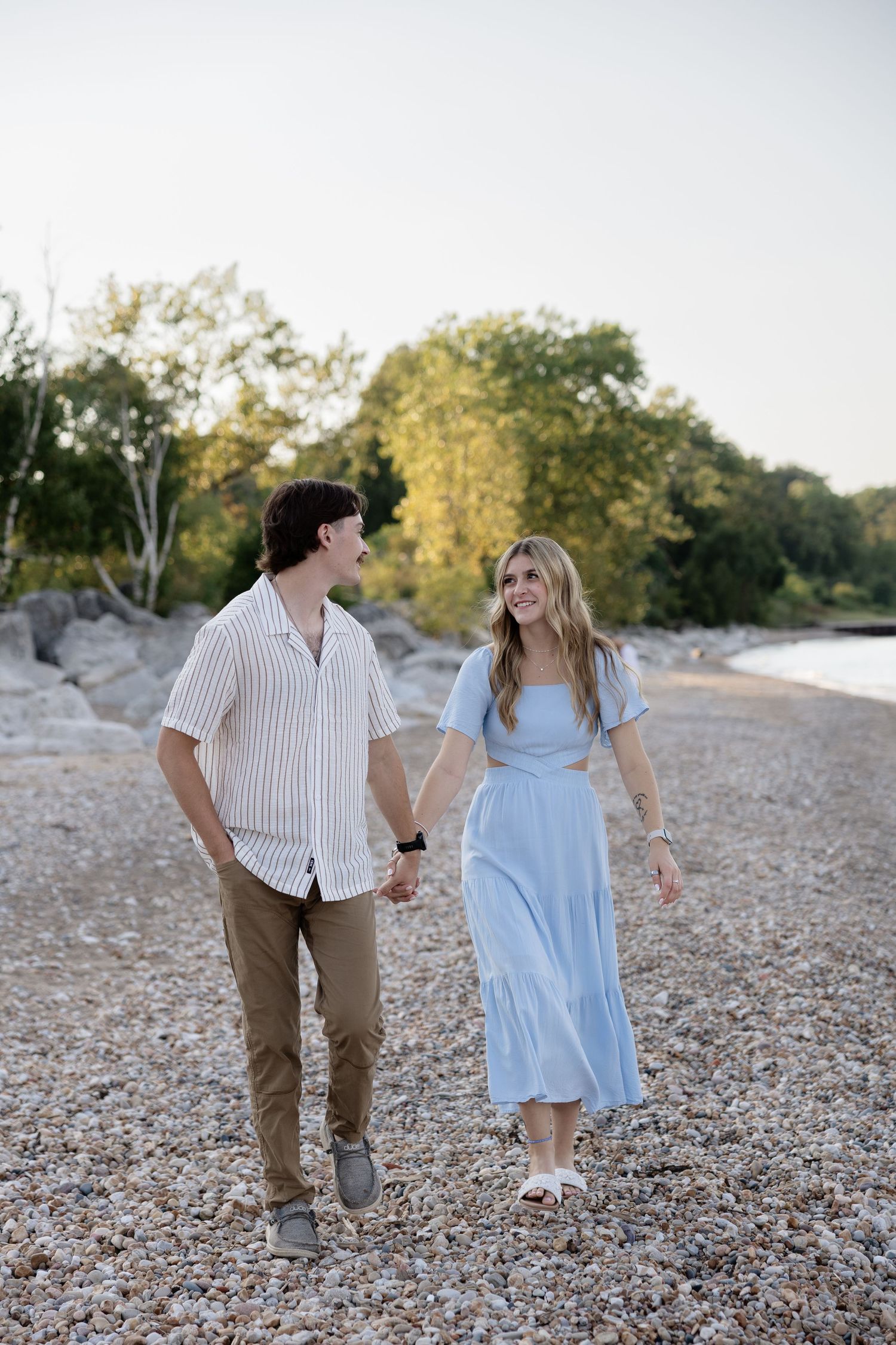 Couple holding hands and walking on a rocky beach shore surrounded by lush green trees at sunset.