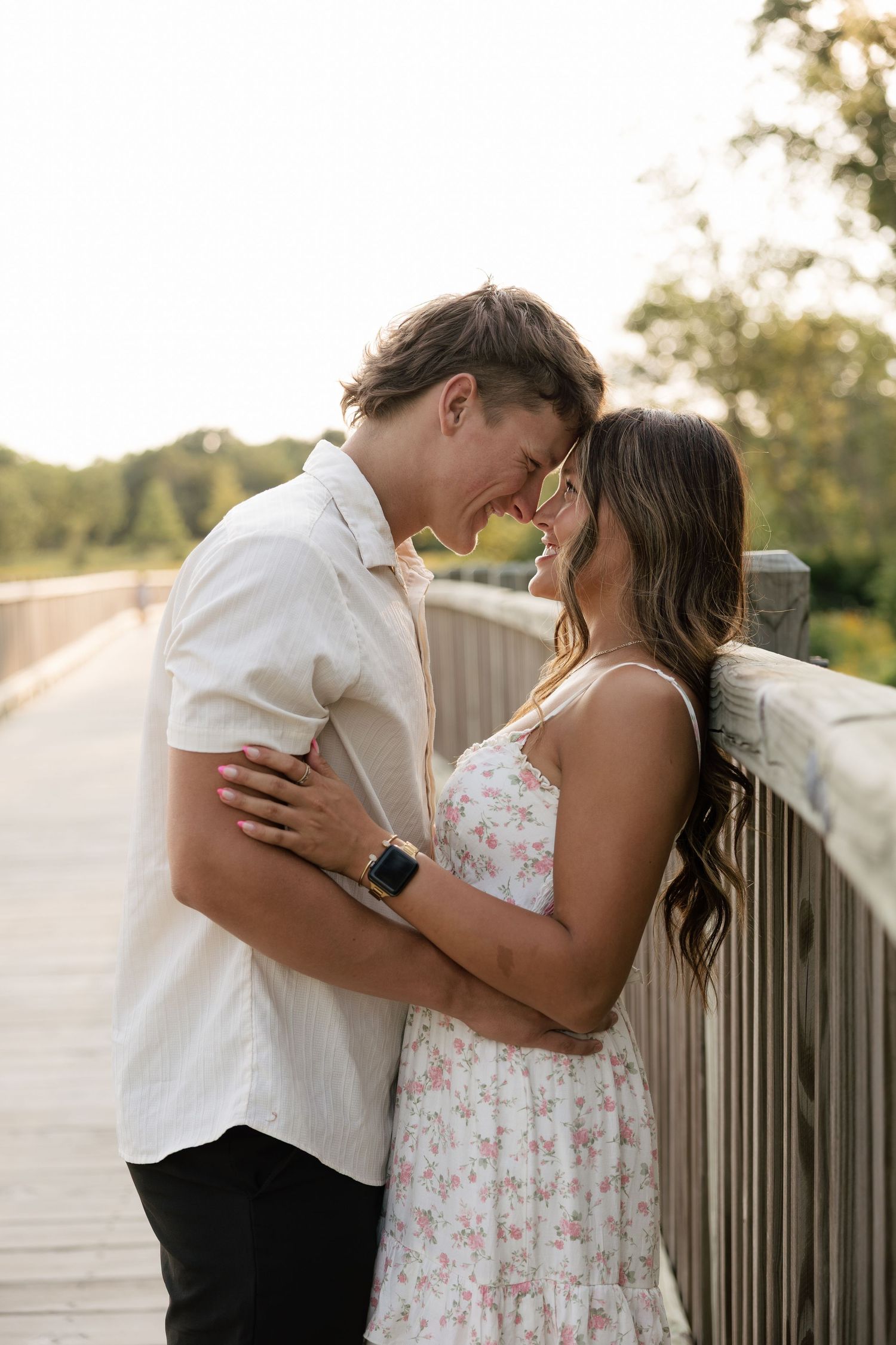 Romantic couple embracing on a wooden bridge at sunset, wearing a floral dress and white shirt.
