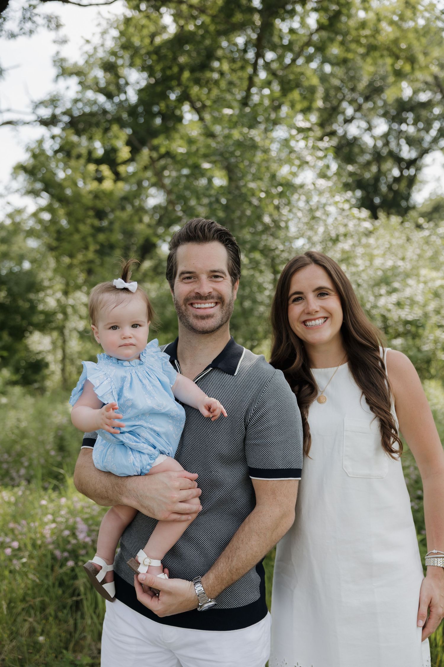 Smiling couple holding their baby girl in a lush outdoor garden setting.
