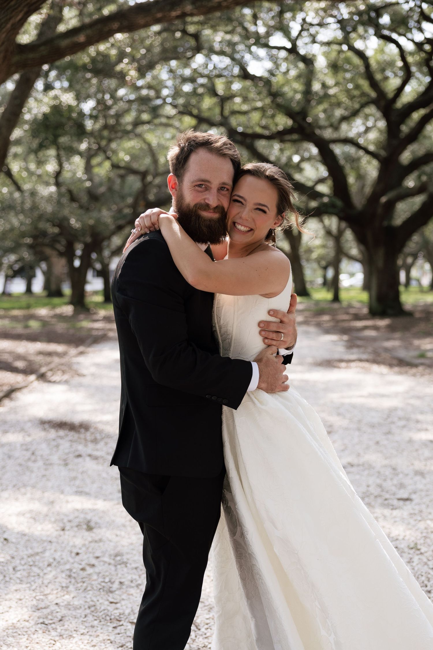 A joyful bride and groom embrace outdoors under a canopy of oak trees on their wedding day.