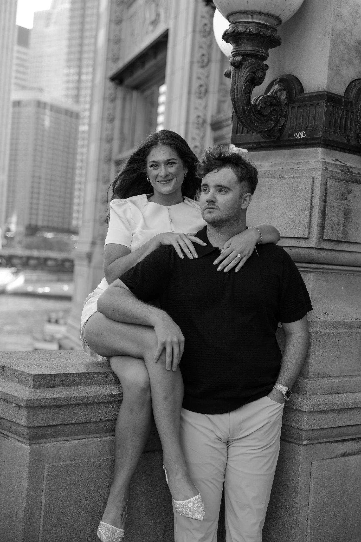 Black and white photo of a couple posing outdoors, woman in white dress sitting on ledge with arm around man in black tee.