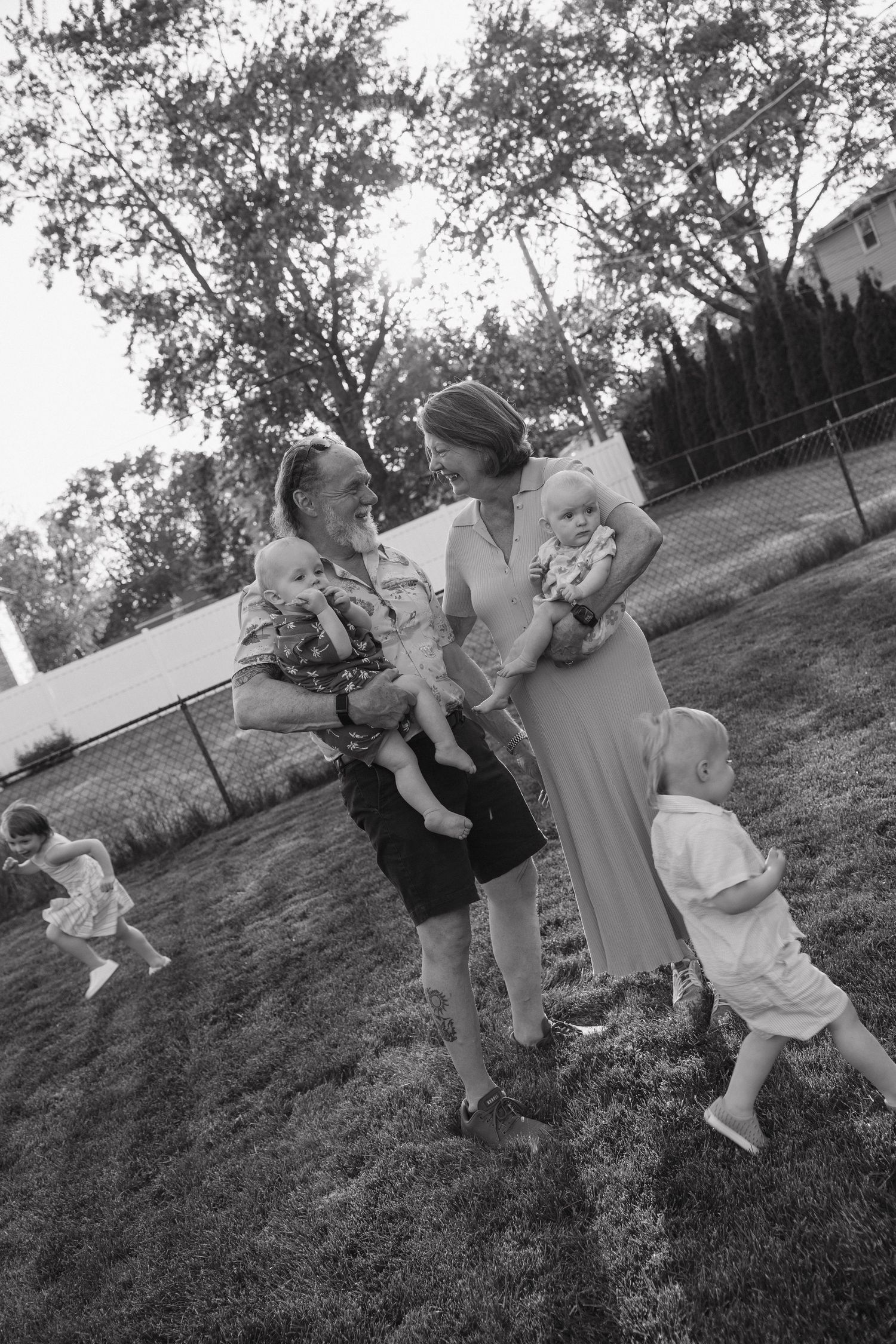 Black and white photo of grandparents holding babies with toddlers playing in backyard.