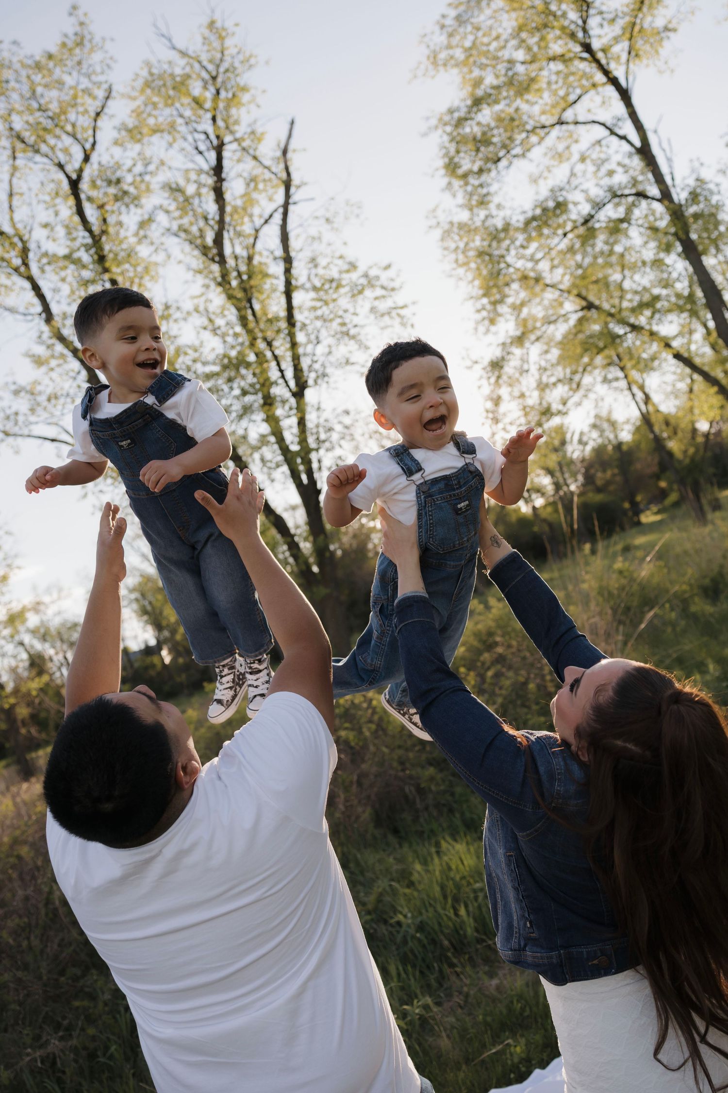 Parents joyfully lifting their twin toddler sons in denim overalls into the air outdoors.
