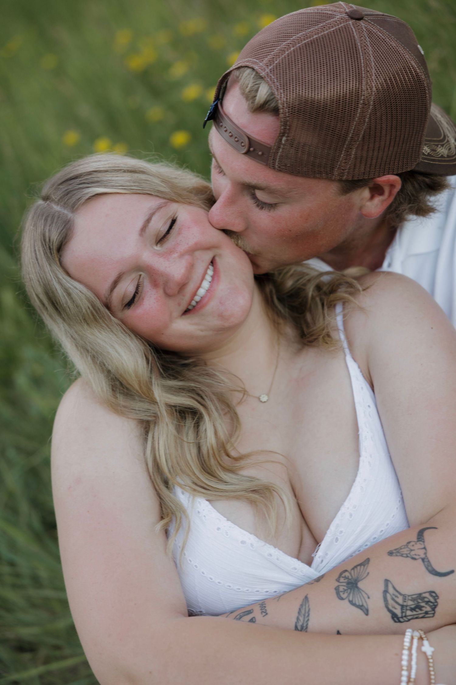 Man in brown cap kissing smiling tattooed blonde woman on cheek in a sunny green meadow.
