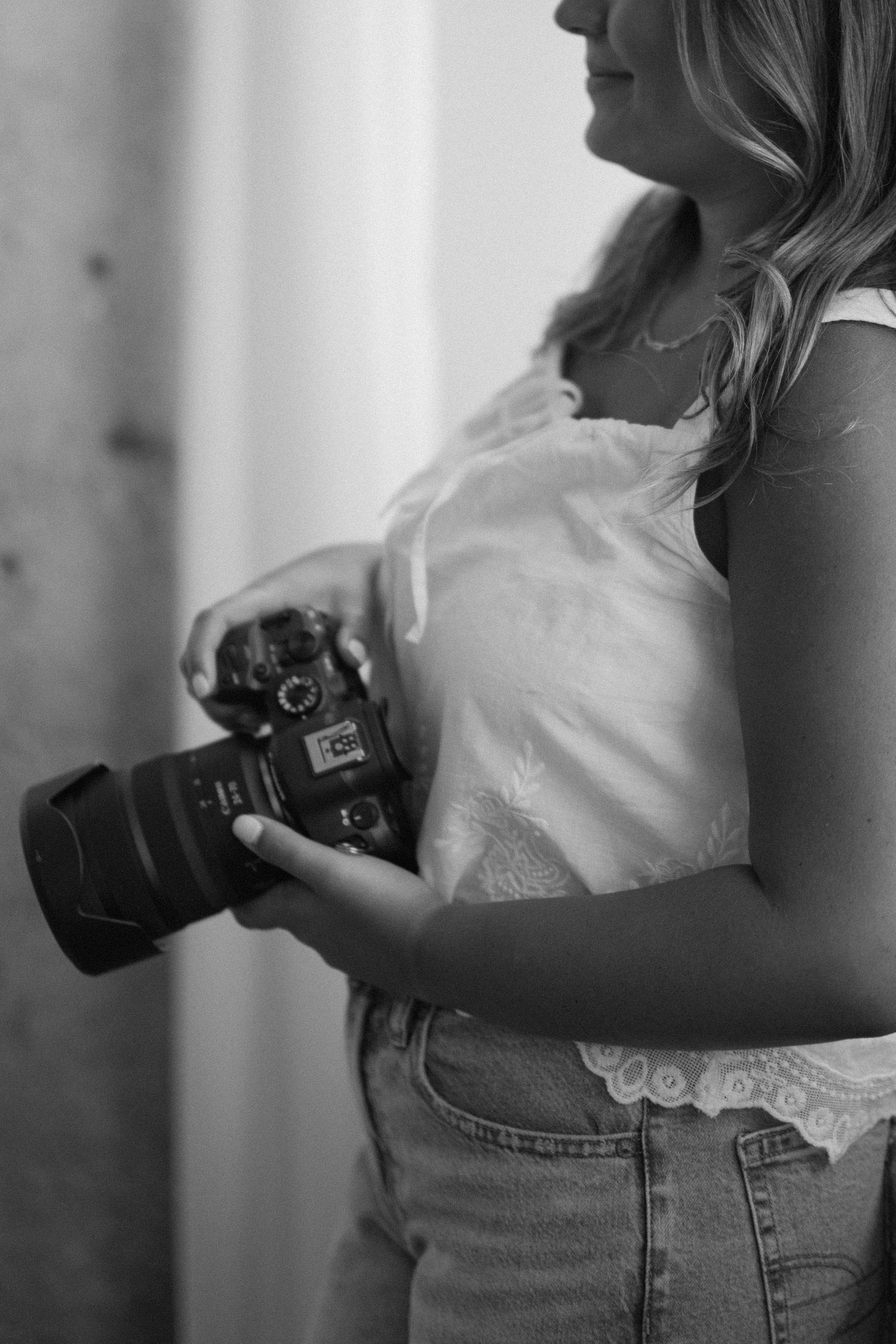 Black and white photo of a woman holding a DSLR camera, wearing a white lace top and jeans.