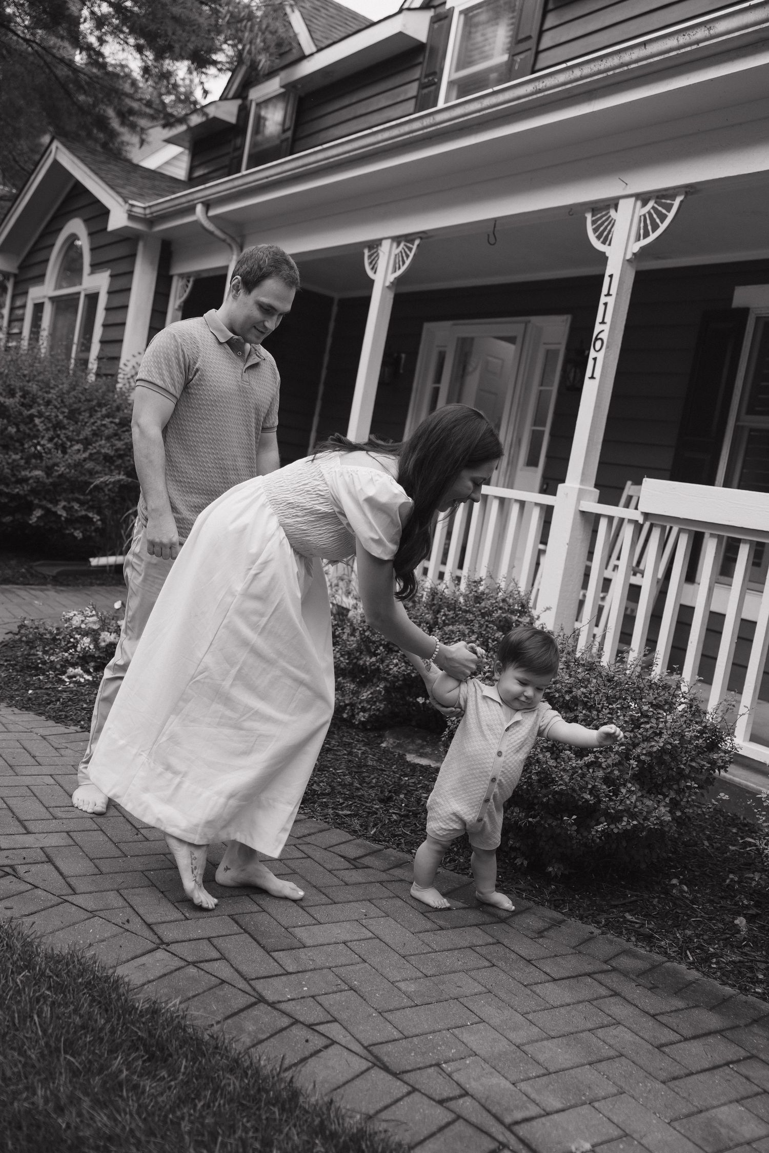 Black and white photo of parents helping toddler walk on brick path in front of charming suburban home.