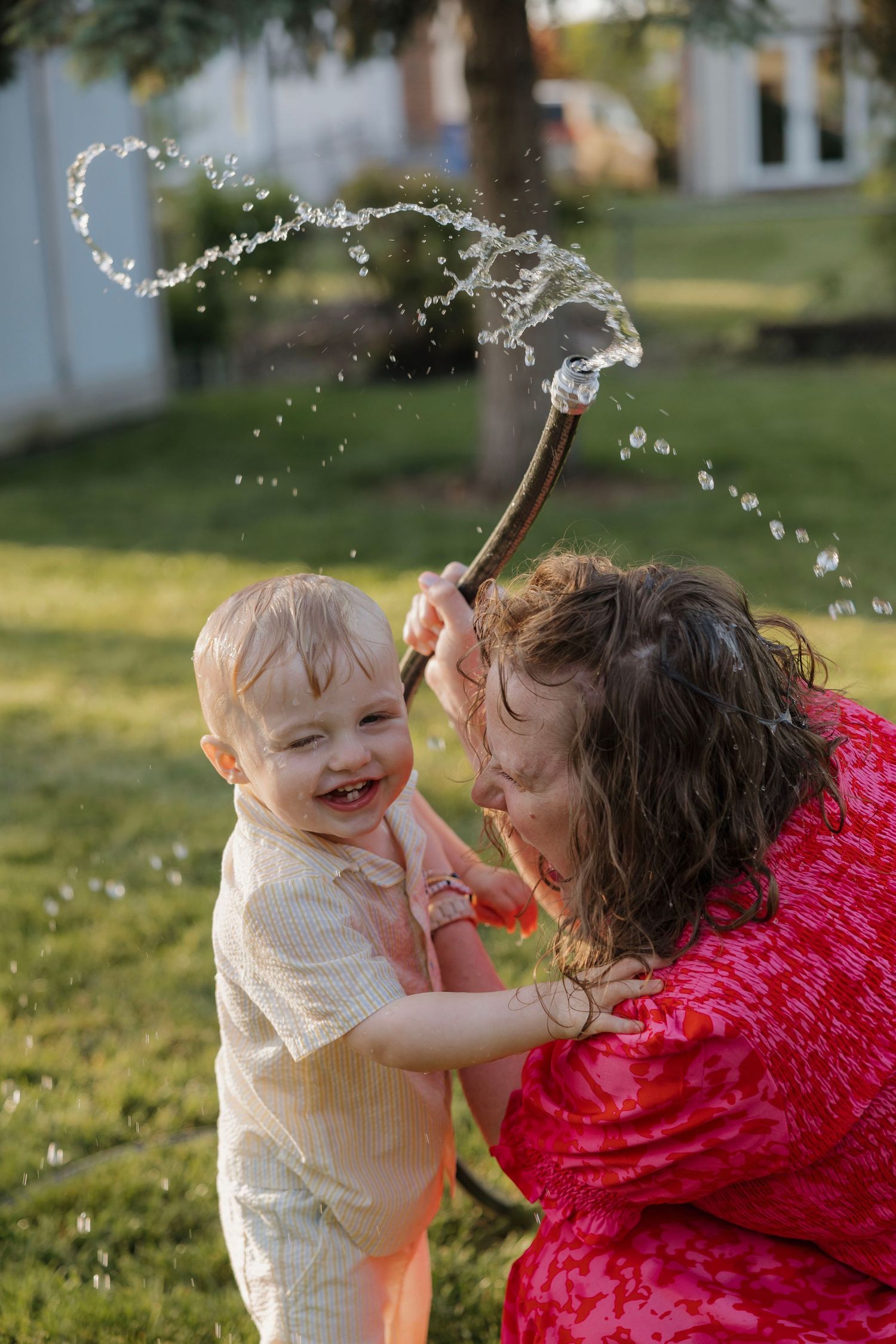 Wet toddler and curly-haired woman laughing while playing with garden hose outdoors.