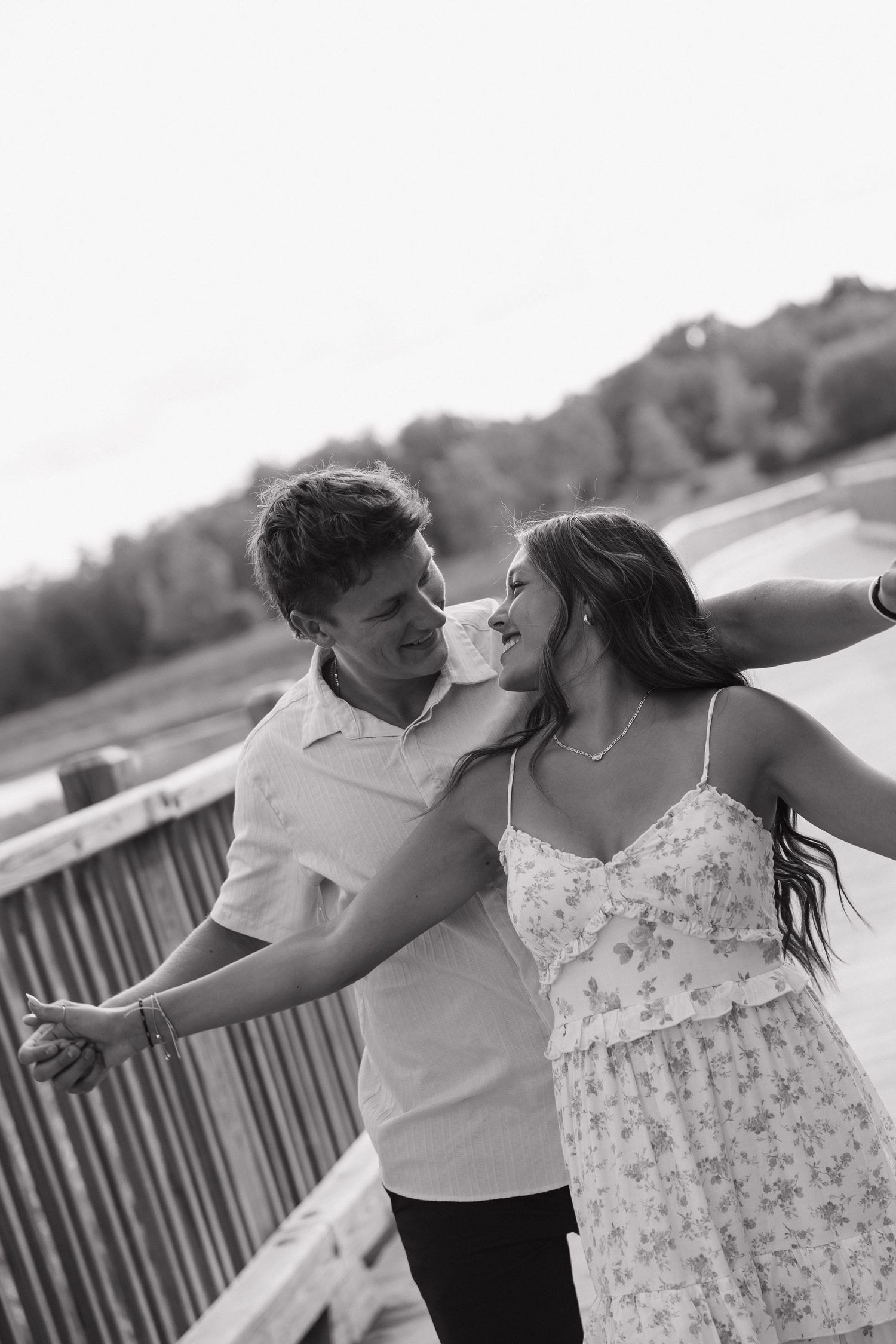 Black and white photo of laughing couple dancing together on a wooden bridge near a lake.