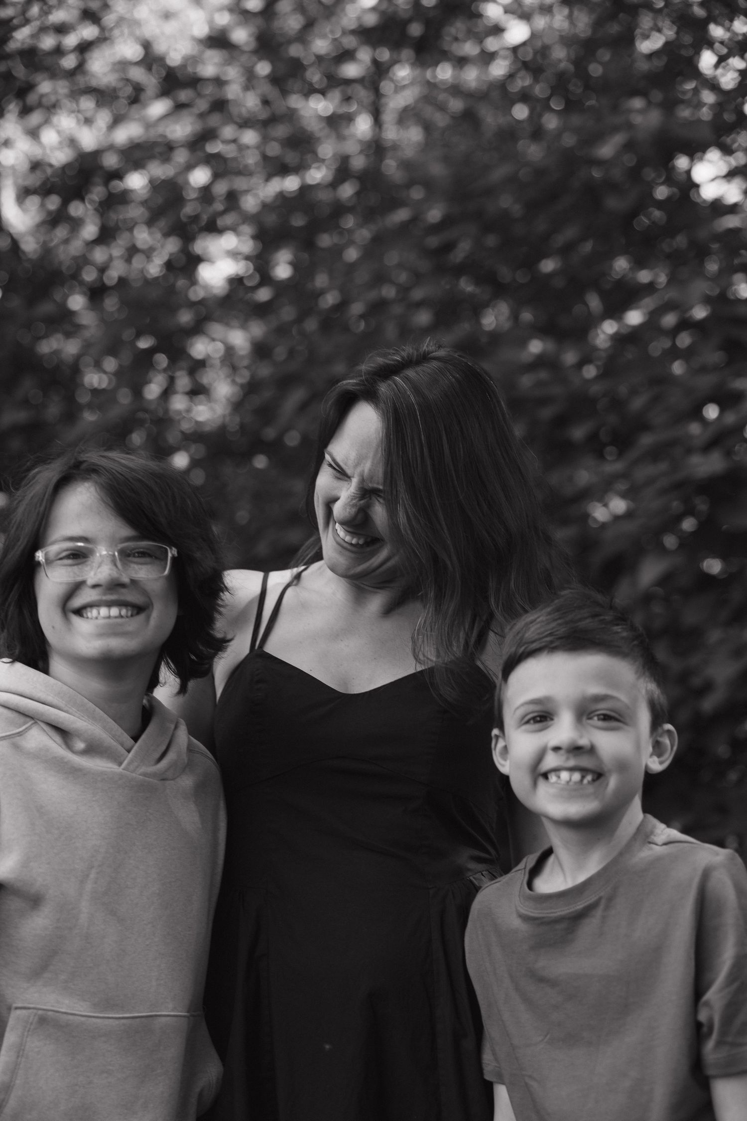 Black and white photo of a smiling mother with her two children outdoors among trees.