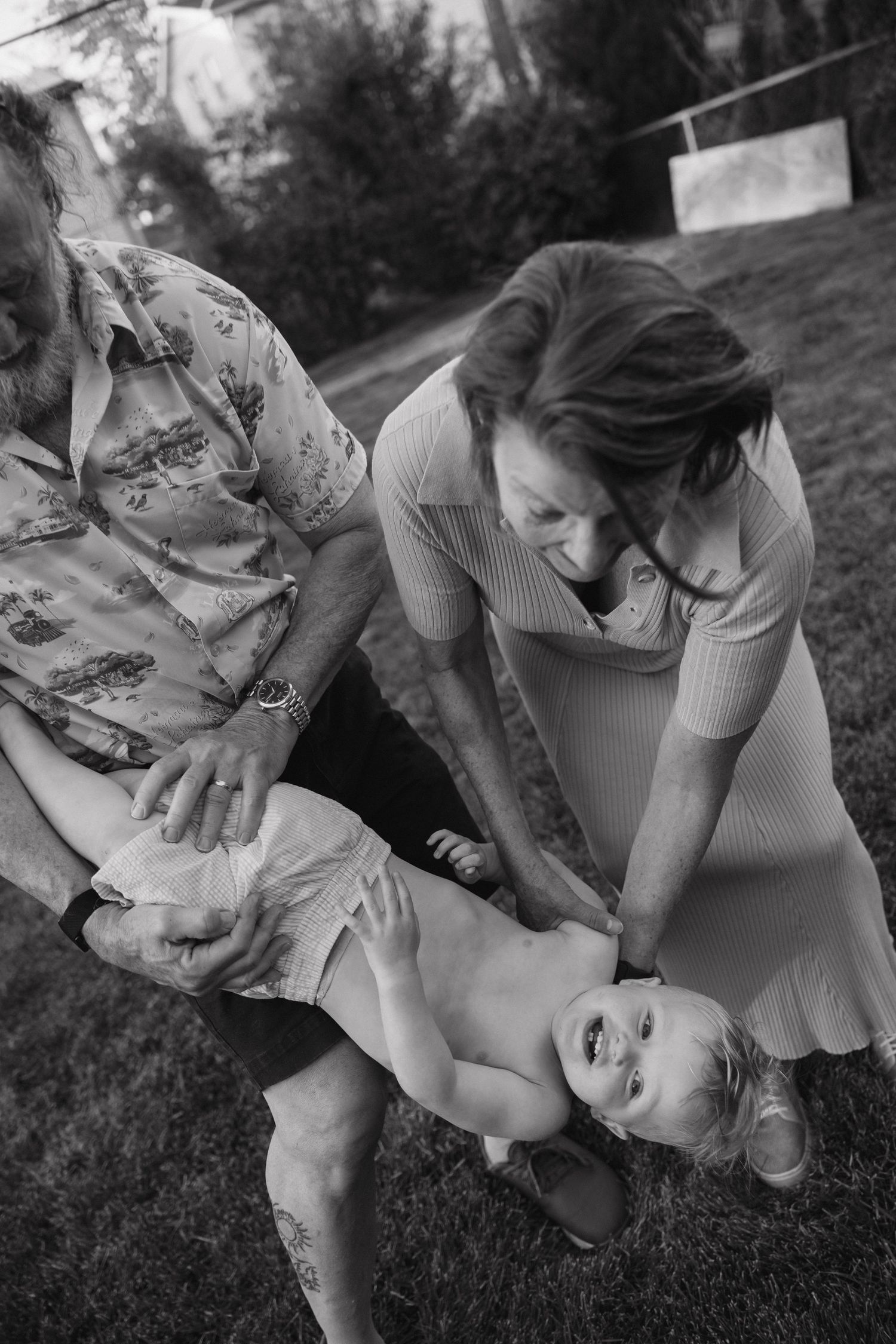 Black and white photo of grandparents playfully holding giggling baby upside down on lawn.
