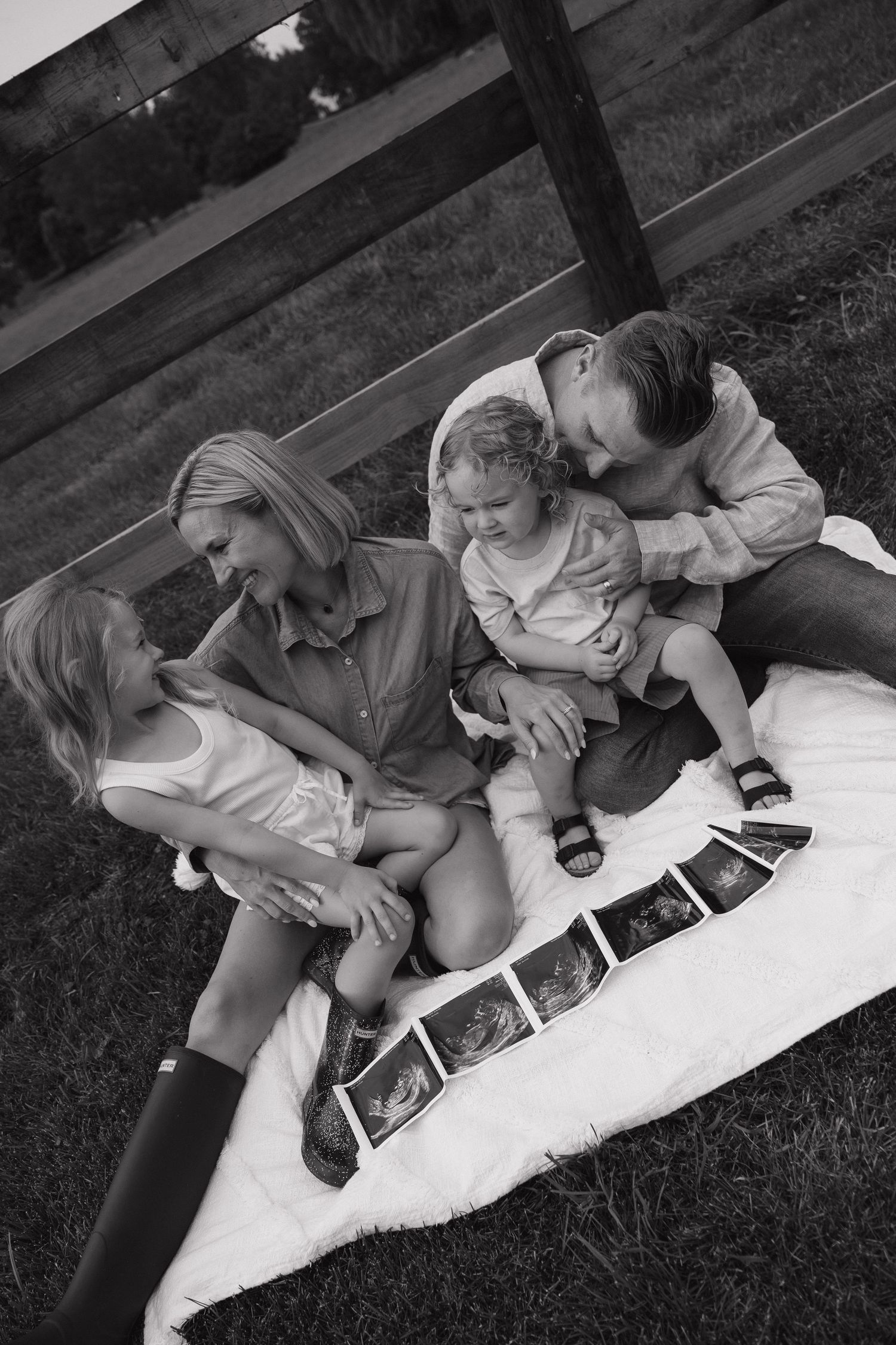 Black and white family photo with parents and two children sitting on blanket with ultrasound photos.