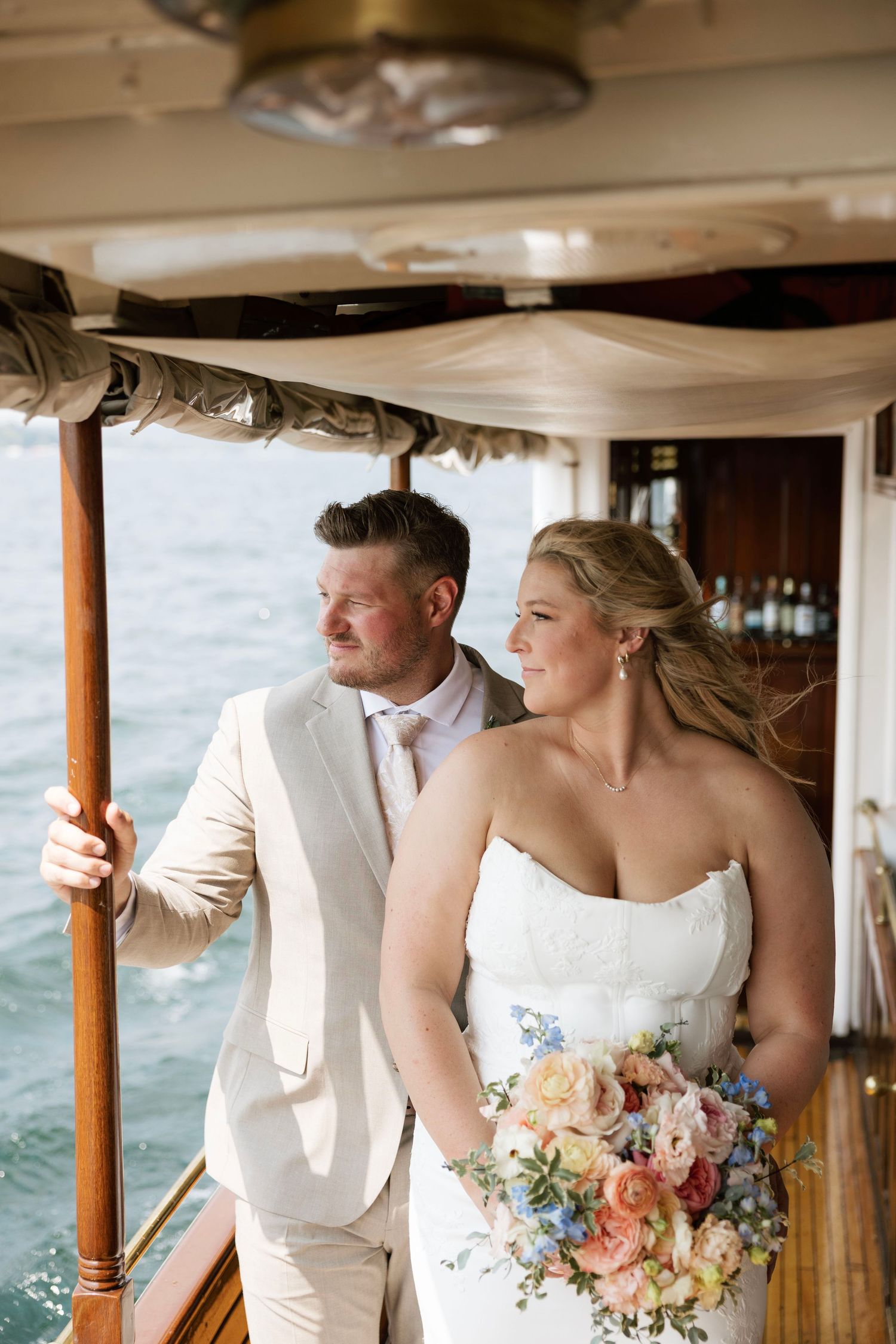 Bride and groom standing on a wooden boat deck, bride holding colorful bouquet with ocean in background.