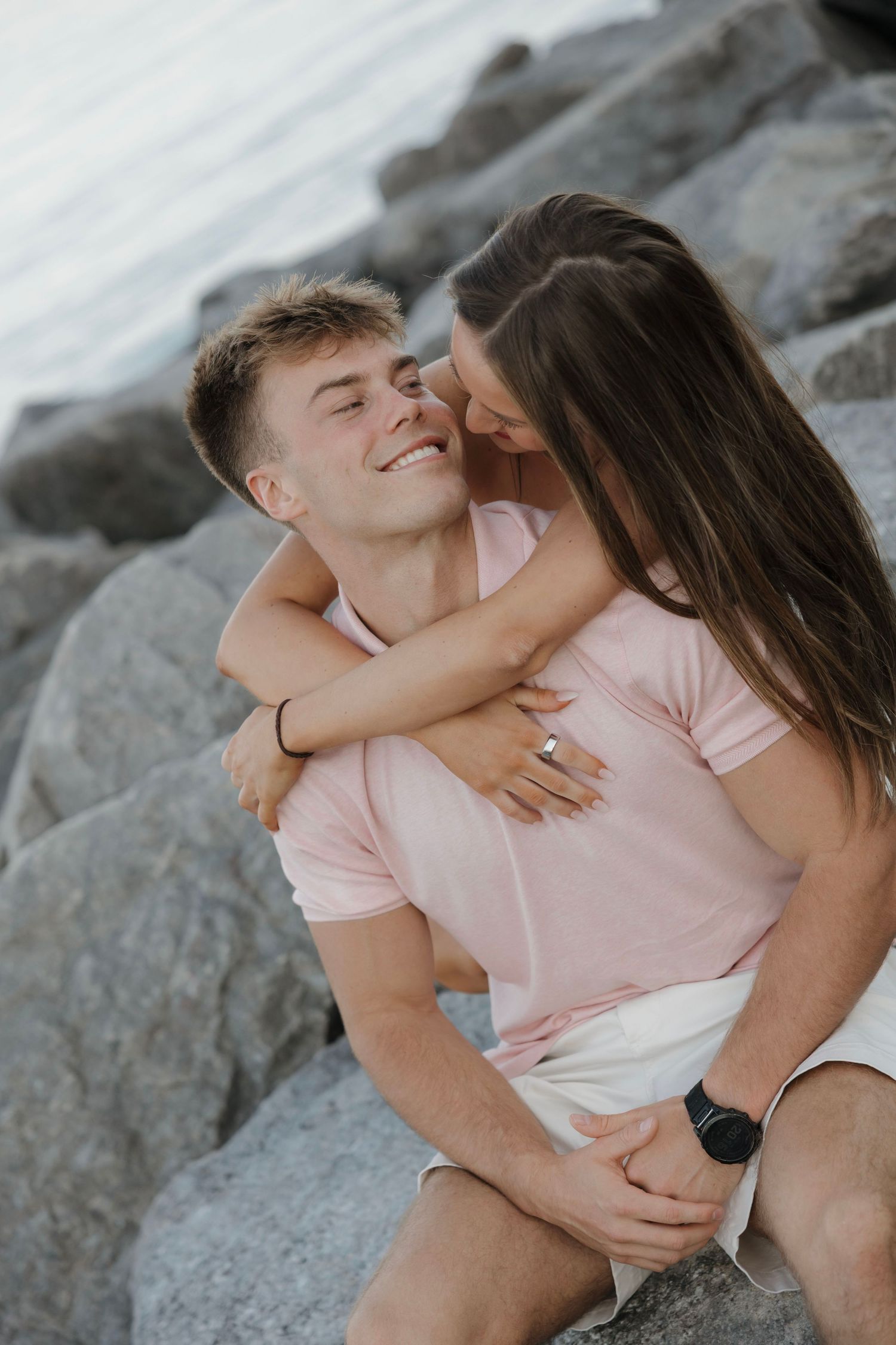Happy couple sitting on rocks near water, woman hugging man from behind as he smiles with eyes closed.