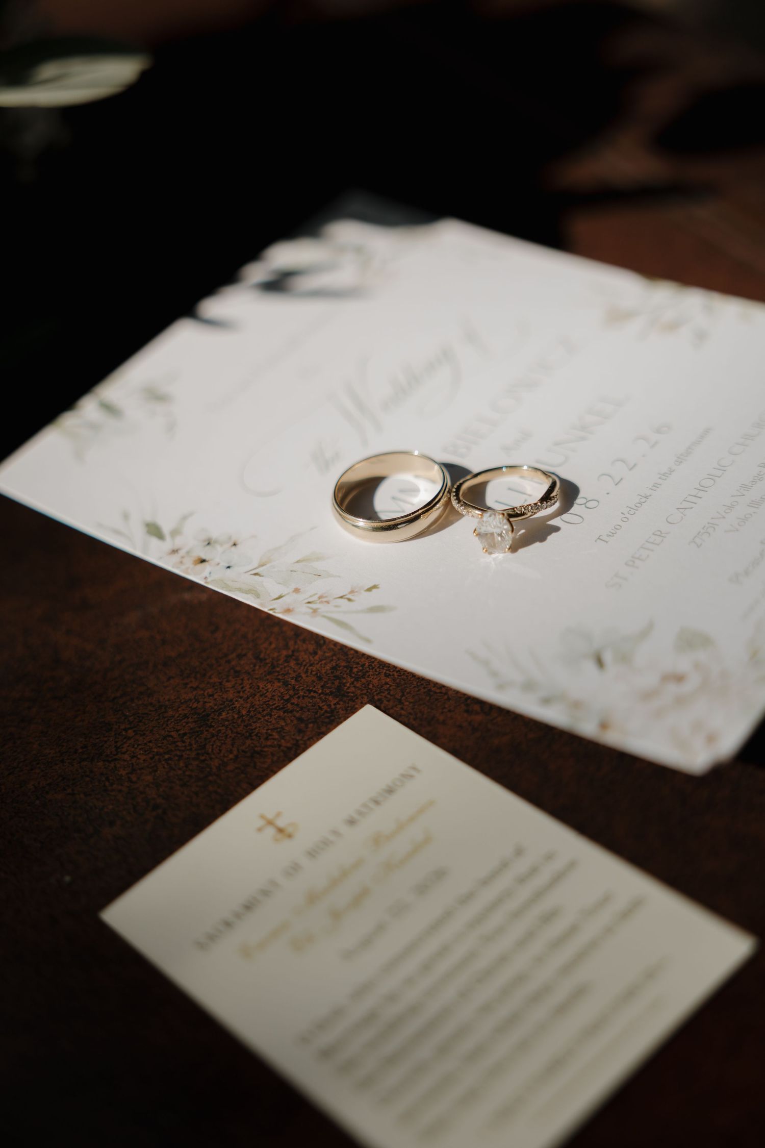 Gold wedding bands and diamond engagement ring resting on elegant floral wedding invitation on dark wood table.