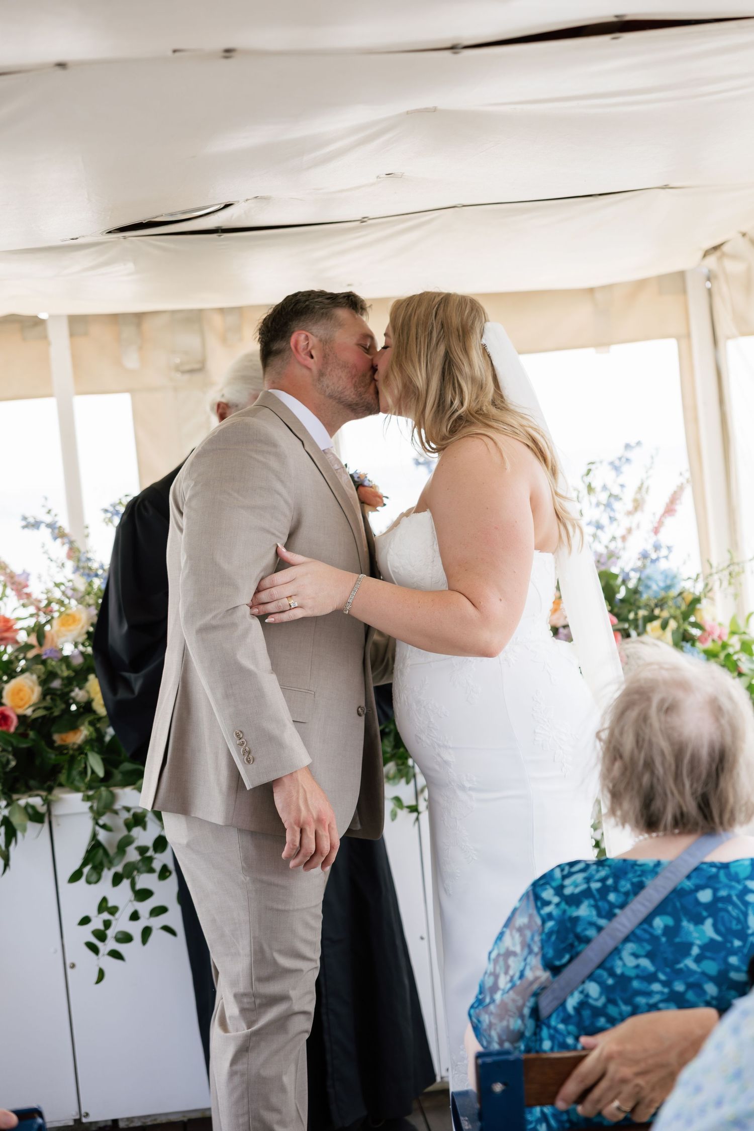 A bride and groom share their first kiss at an outdoor wedding ceremony under a white marquee tent surrounded by colorful flowers.
