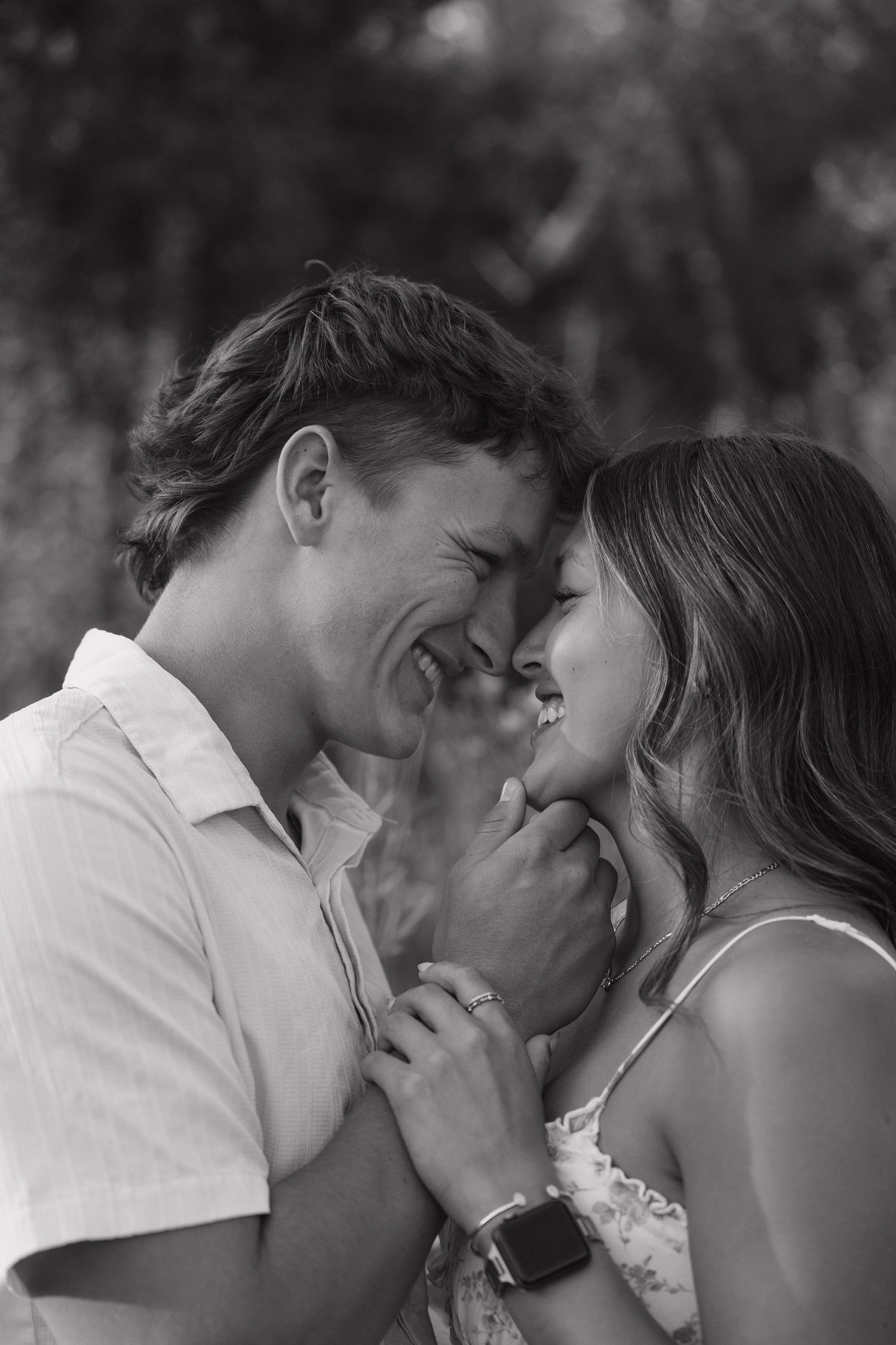 Black and white romantic photo of smiling couple touching foreheads tenderly in an outdoor setting.
