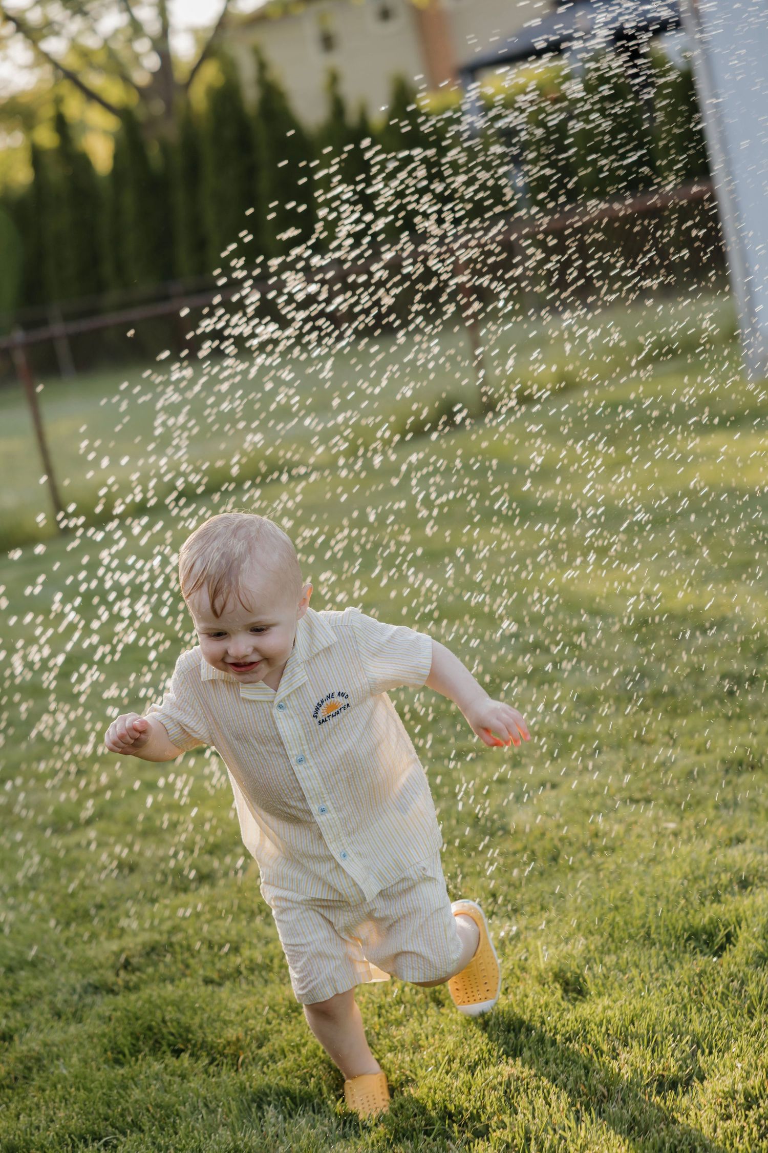 Happy toddler in white romper running through sprinkler spray on green grass.