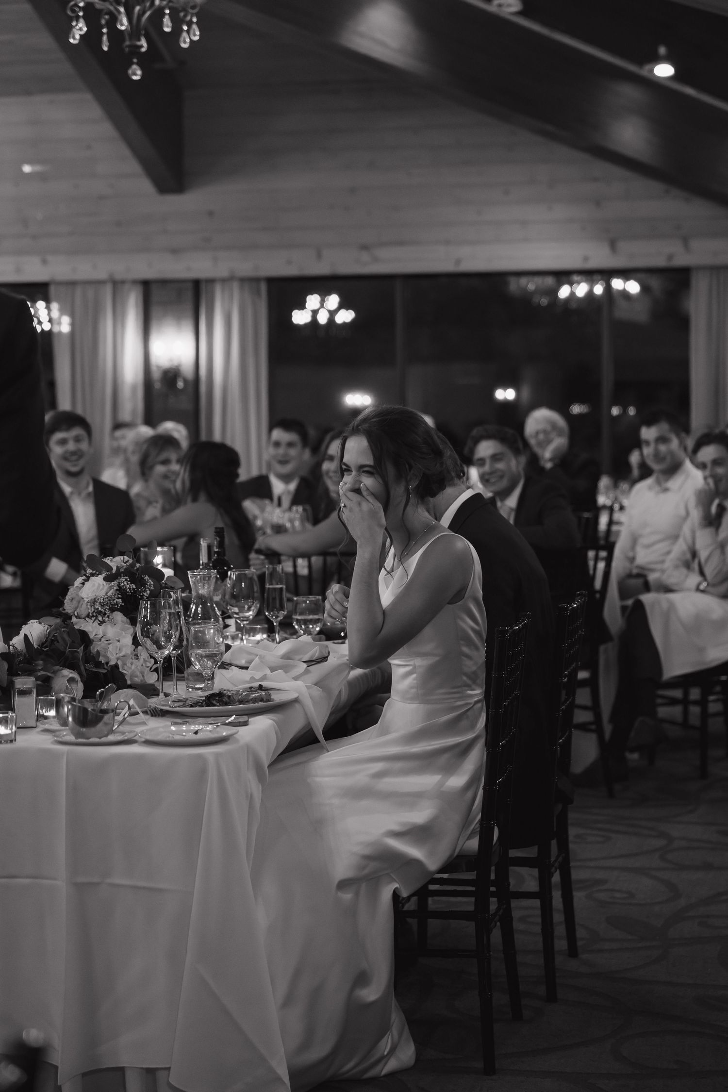 Black and white photo of an emotional bride laughing at her wedding reception dinner table surrounded by guests.