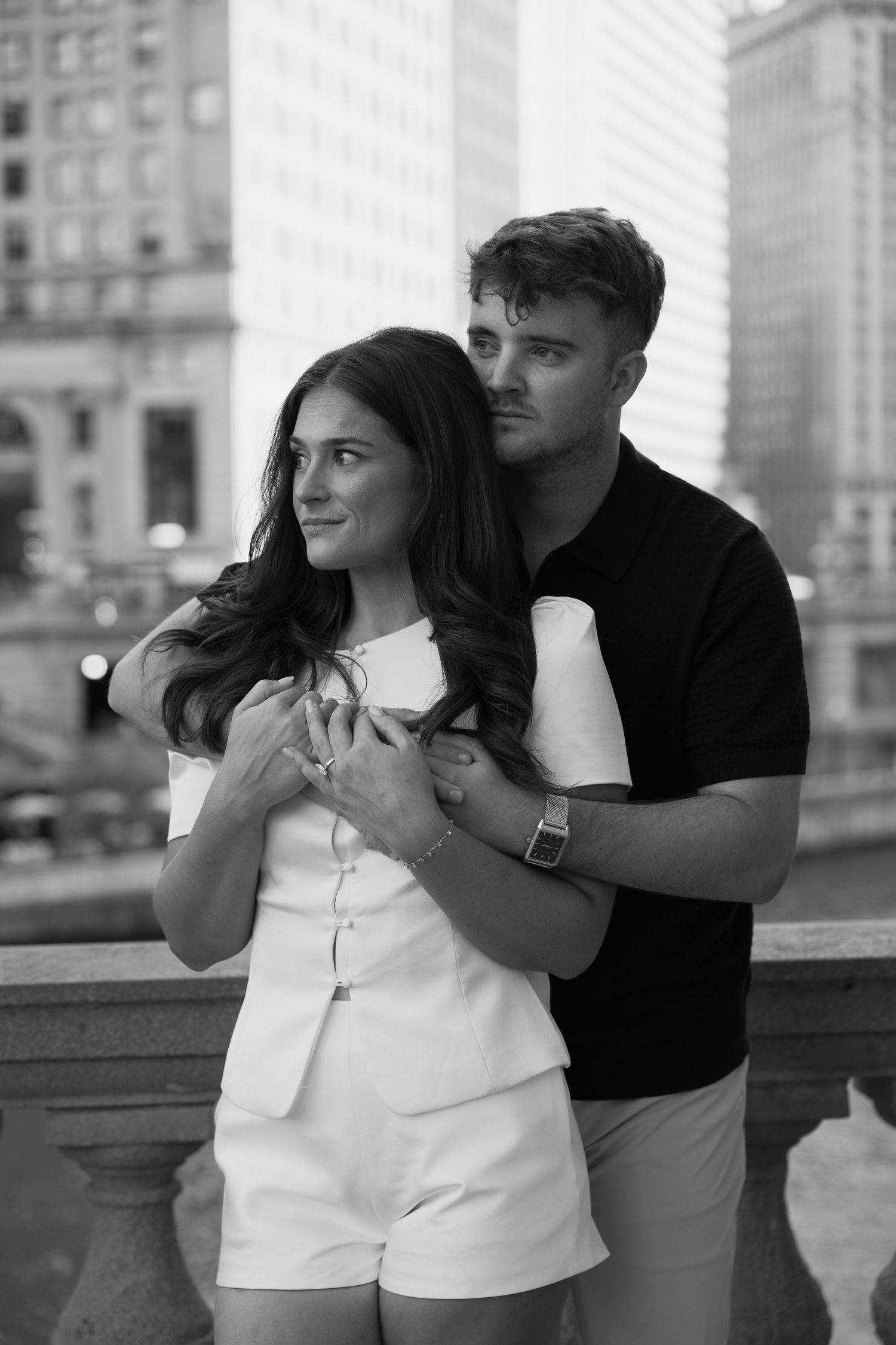 Black and white photo of a couple embracing on a city bridge, woman in white romper, man in black shirt.
