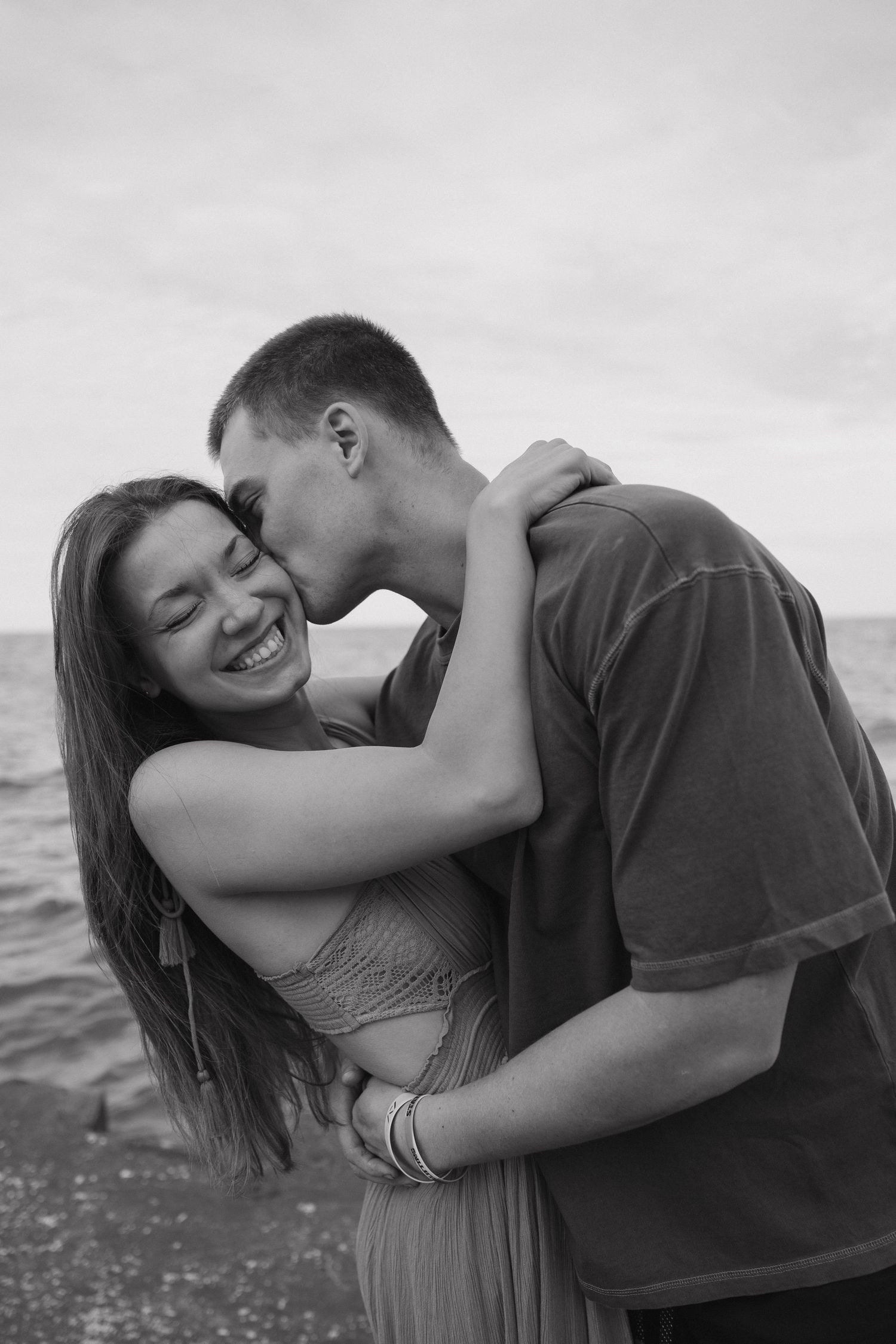 Black and white beach photo of couple embracing and kissing near the ocean shoreline.