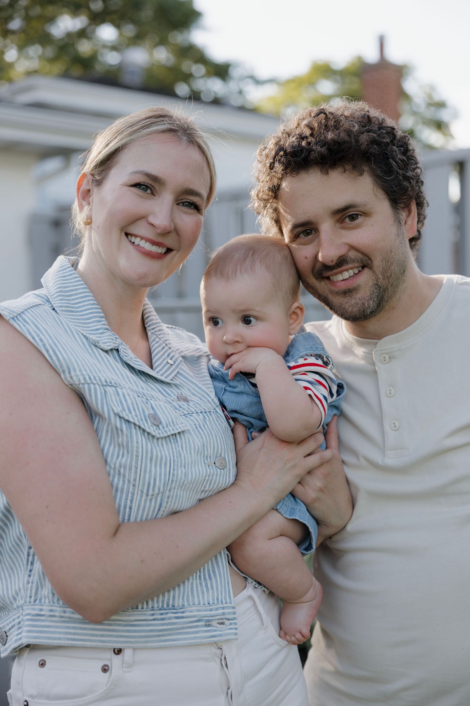Smiling young couple standing outdoors holding their baby dressed in denim overalls and striped shirt.