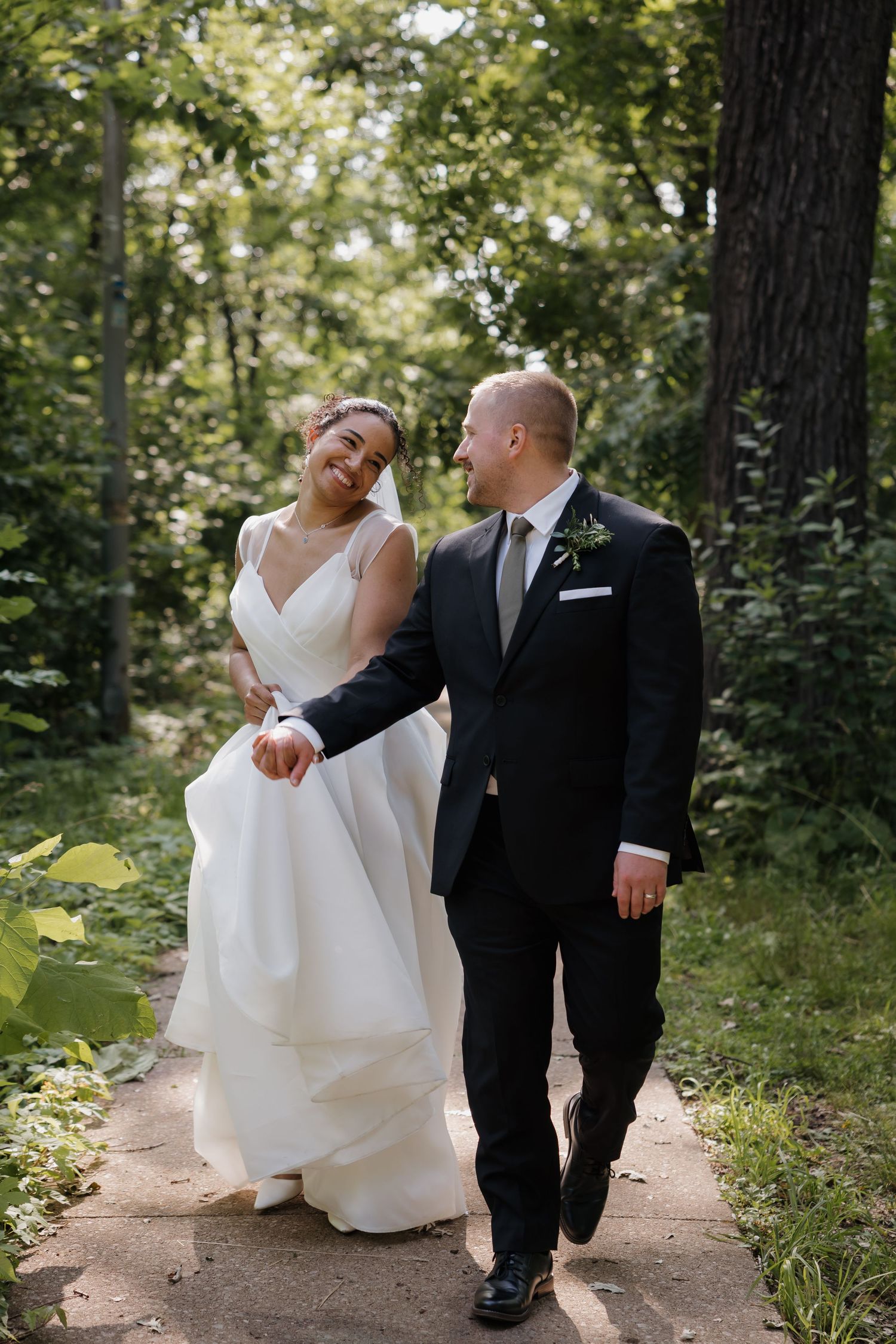 Interracial newlywed couple walking hand in hand on a forest path, bride laughing in layered white gown.