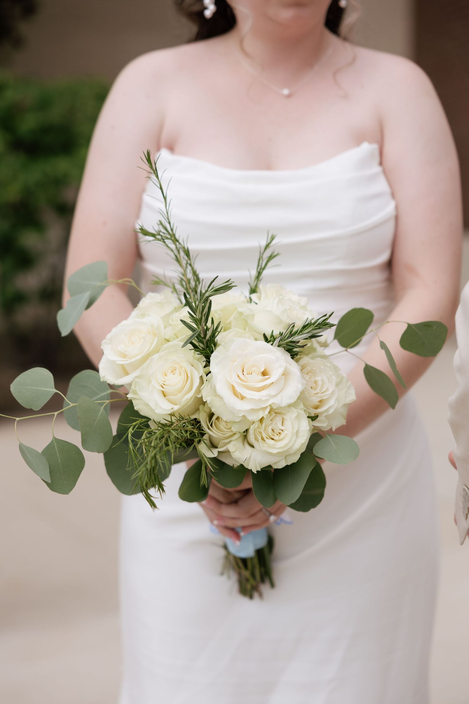 Bride in strapless white gown holding elegant bouquet of white roses and eucalyptus greenery at wedding ceremony.