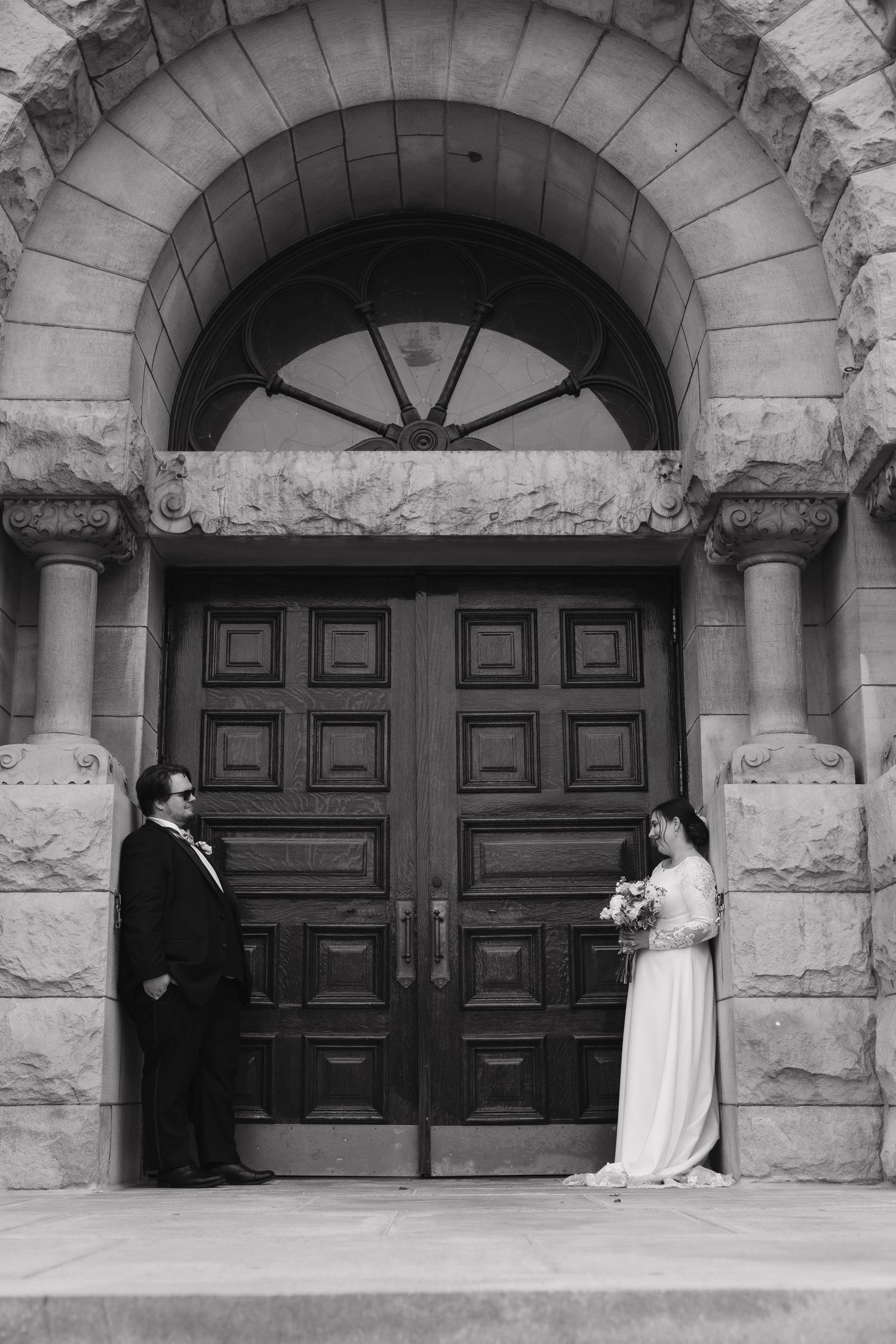 Black and white photo of bride and groom standing apart at ornate church doors with stone arch.