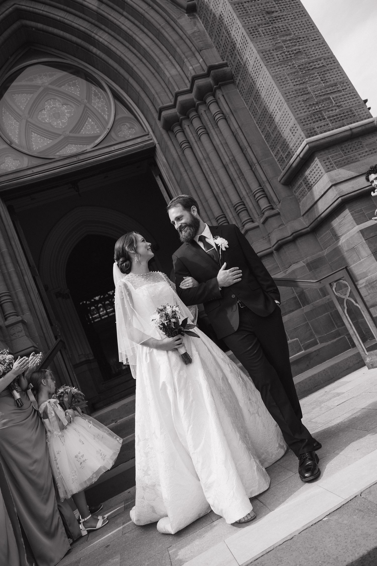 Black and white photo of a smiling bride and groom exiting a stone church, with flower girls nearby.