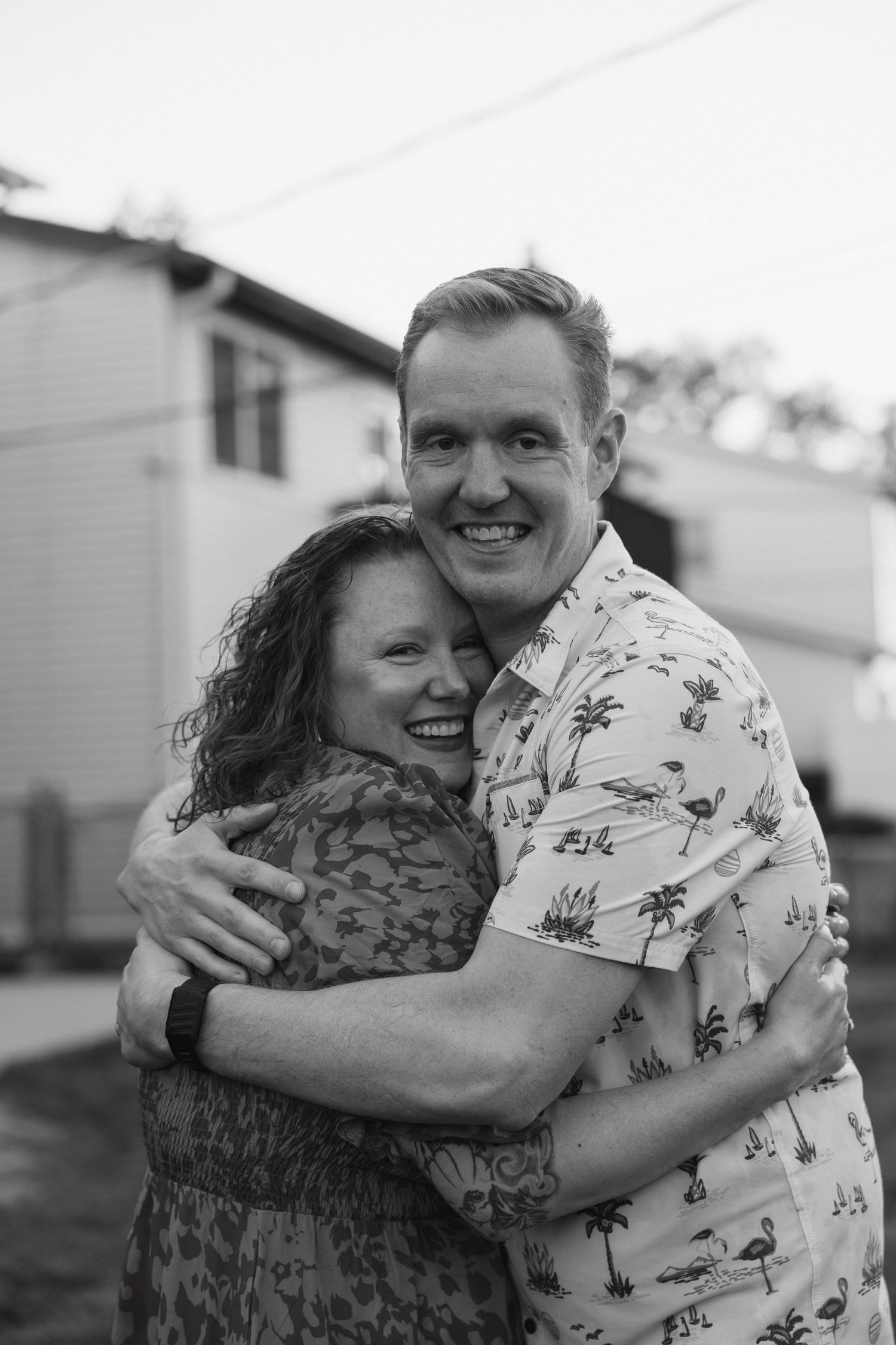 Black and white portrait of smiling couple embracing outdoors near white building.