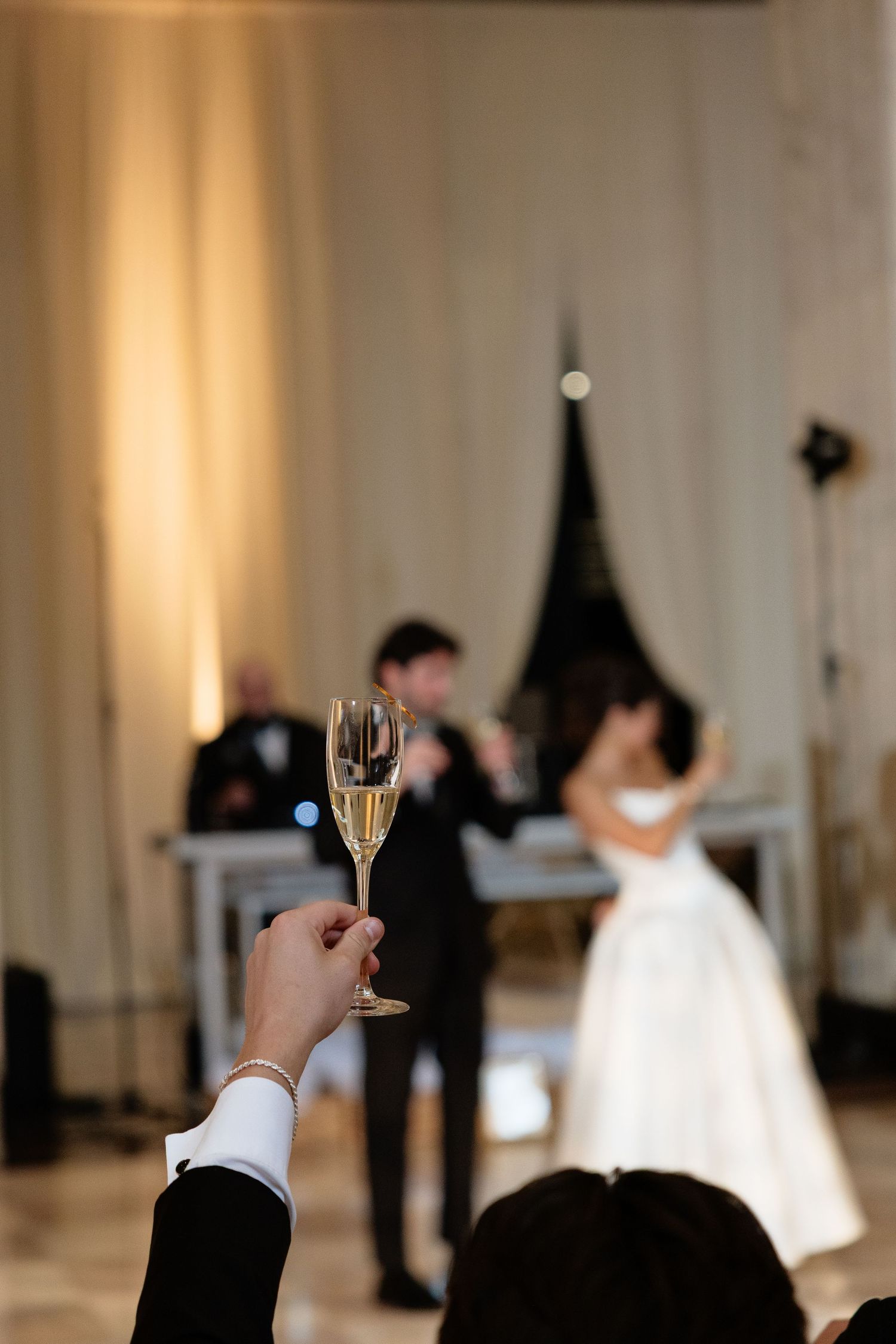 A guest raises a champagne flute during a wedding reception toast while the bride and groom stand in the background.