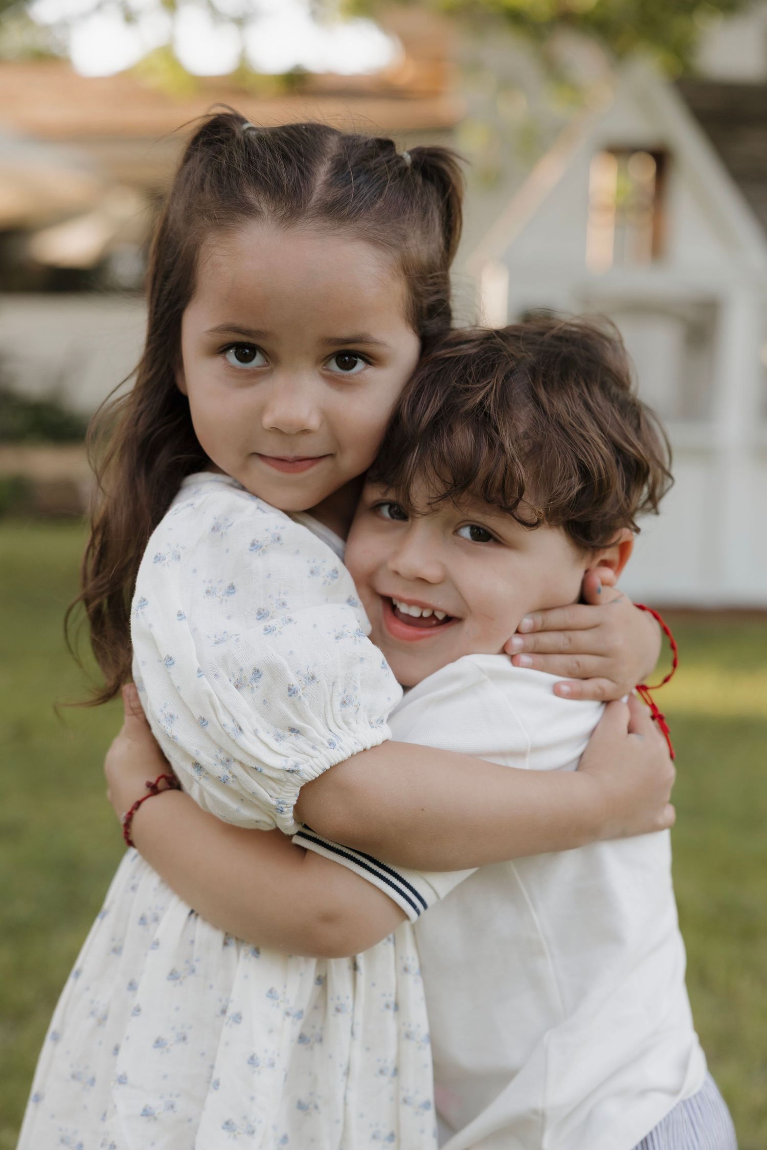 Adorable young girl hugging her little brother in a garden near a white house.