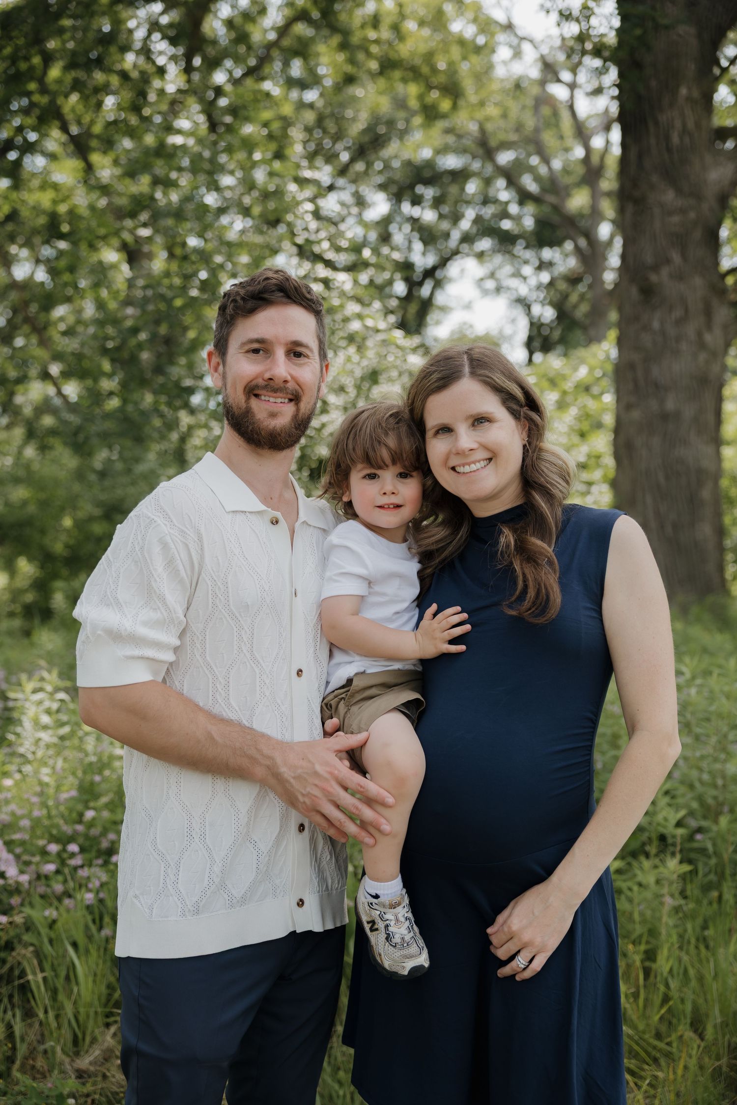 Pregnant woman in navy dress poses with husband and young son in a garden.