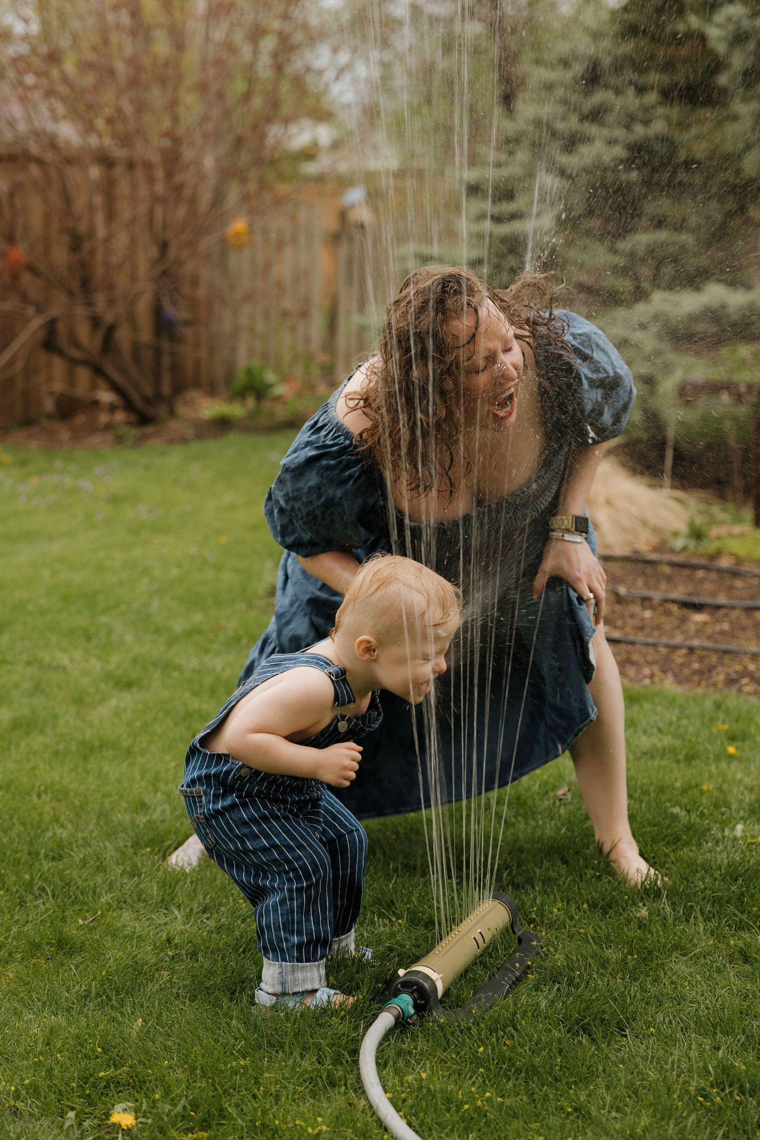 Mother and toddler playing and laughing in backyard sprinkler on green lawn.