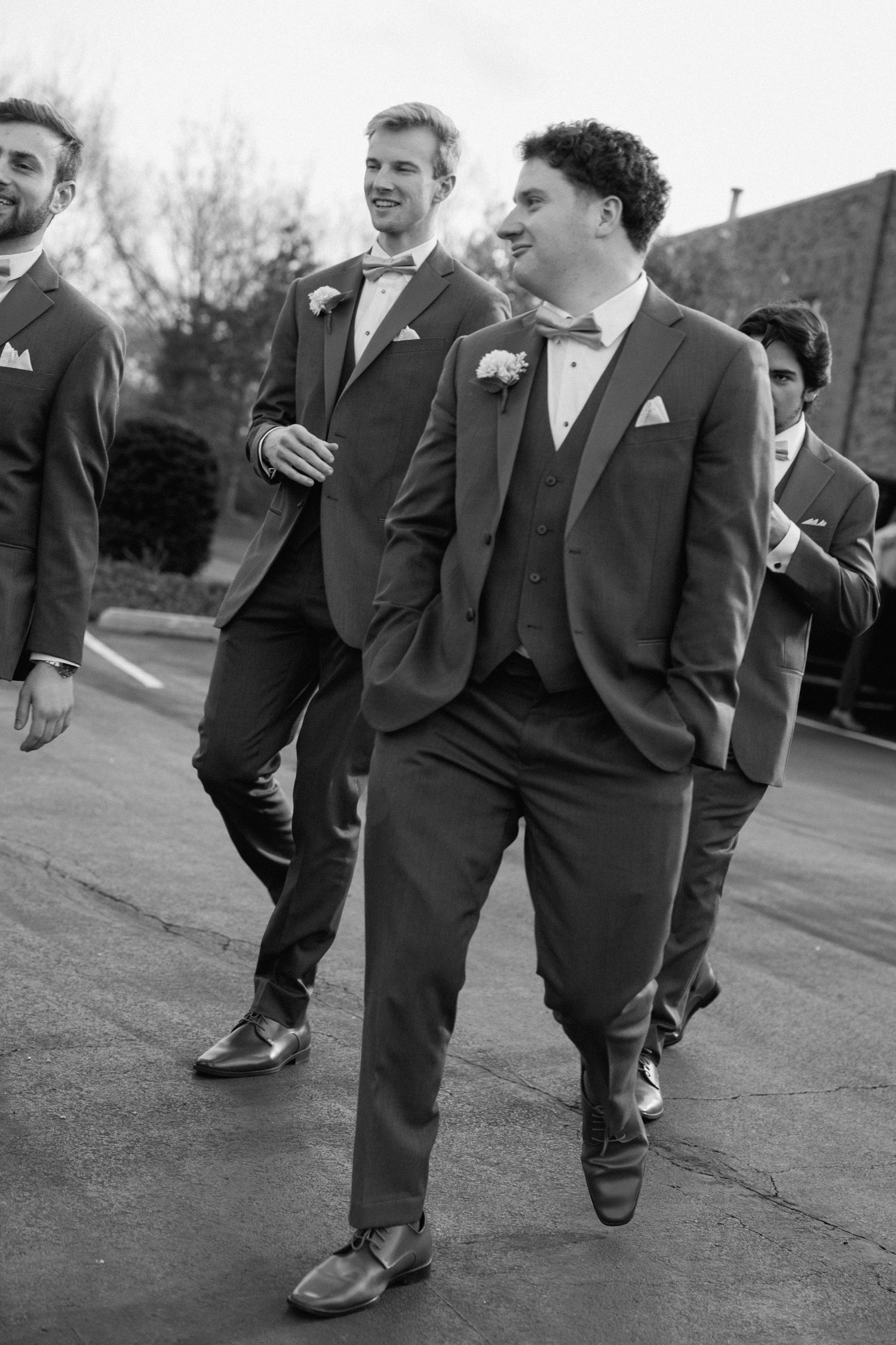 Groomsmen in matching gray suits and bow ties walking together outdoors in black and white wedding photo.