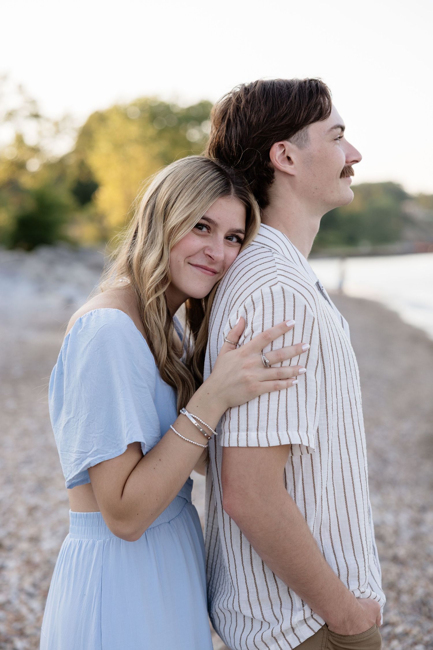 A couple embraces on a rocky beach, woman in blue dress smiling at camera, man in striped shirt looking away.