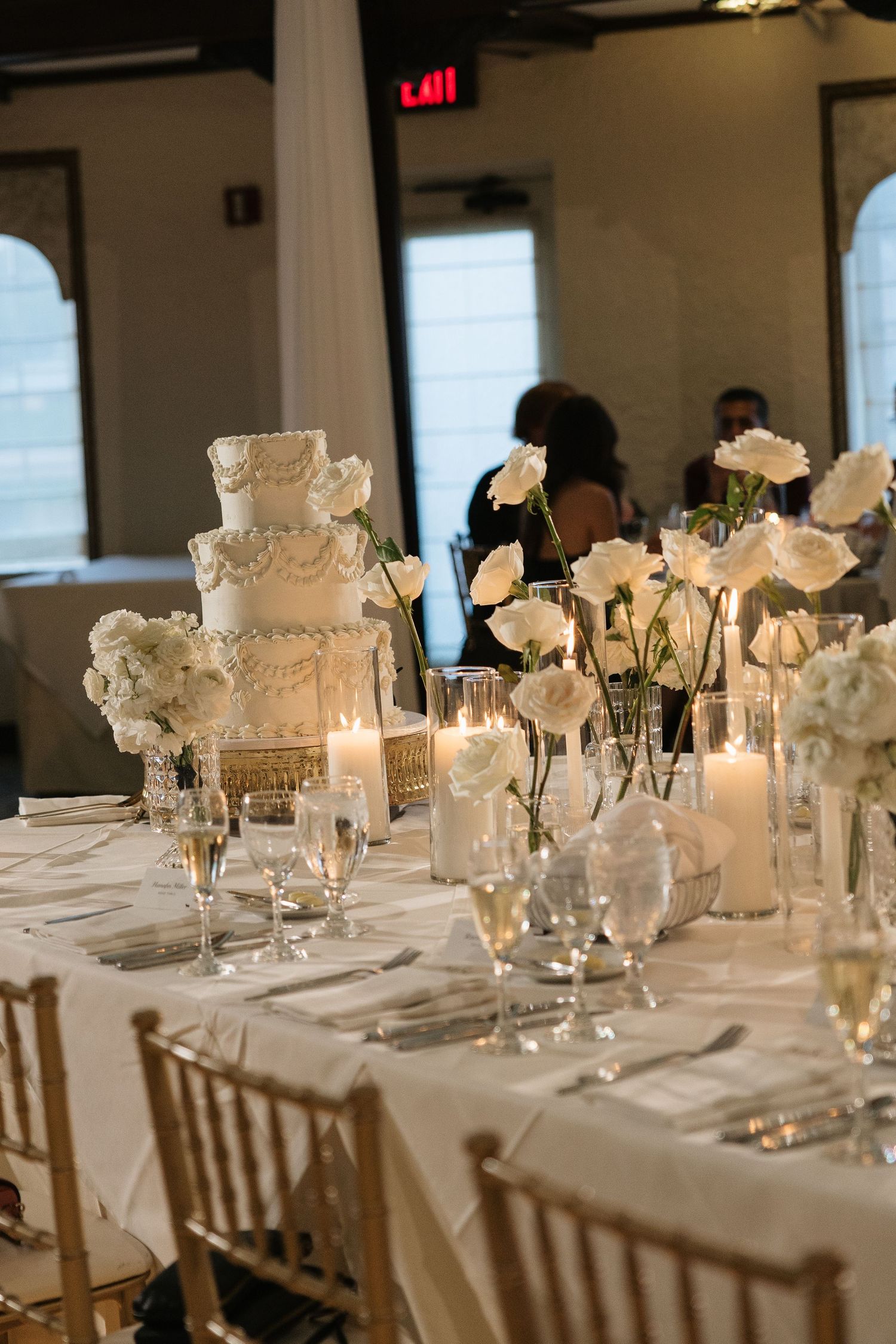 Elegant wedding reception table with white tiered cake, white roses, candles, and crystal glassware on white linen.
