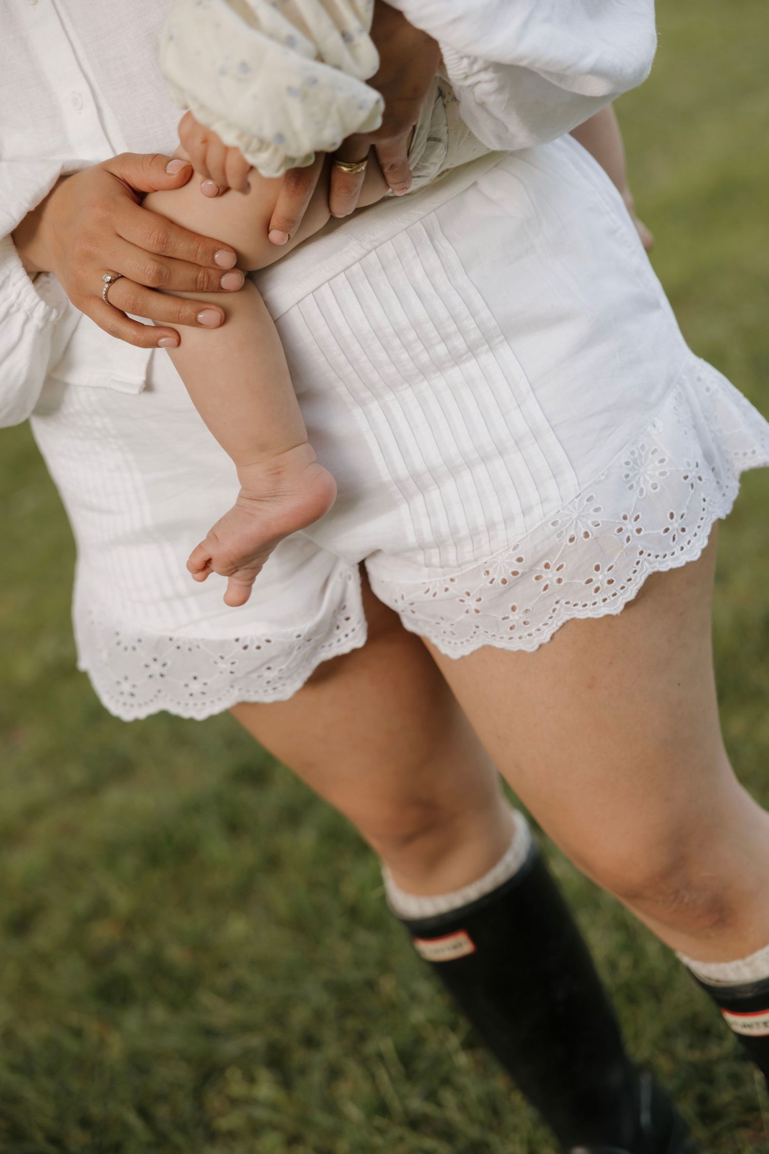 Mother in white eyelet shorts holding newborn baby outdoors in garden.