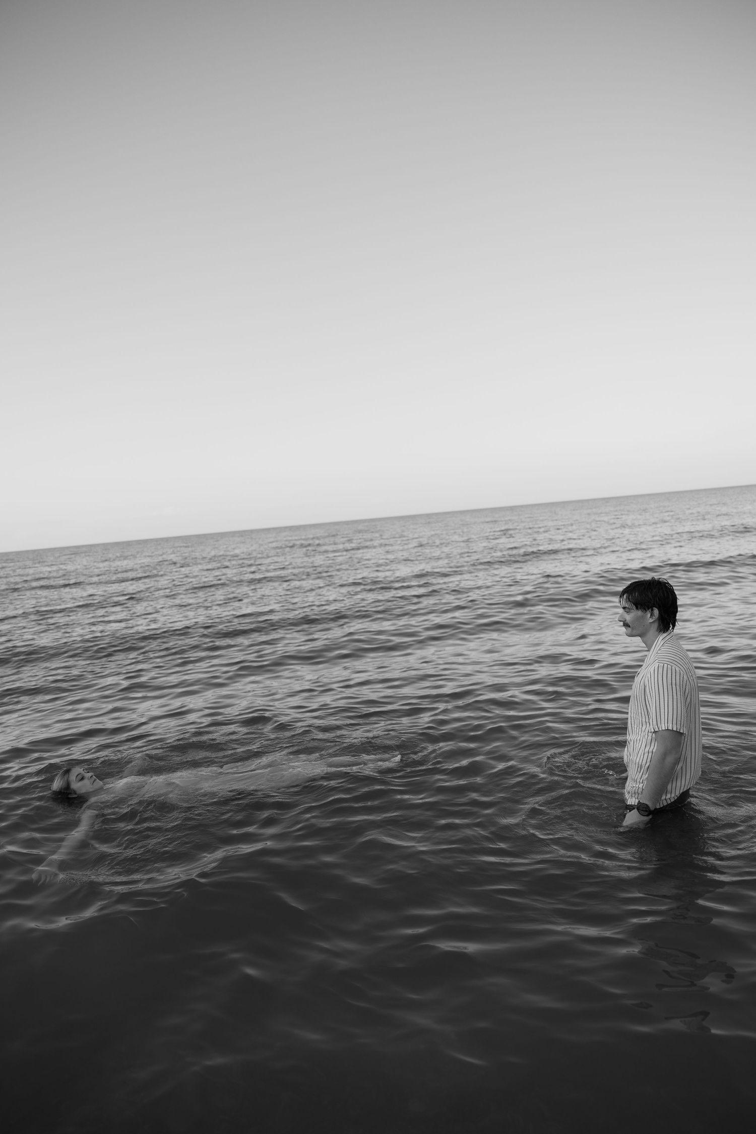 Black and white photo of a person standing waist-deep in calm ocean water at dusk.